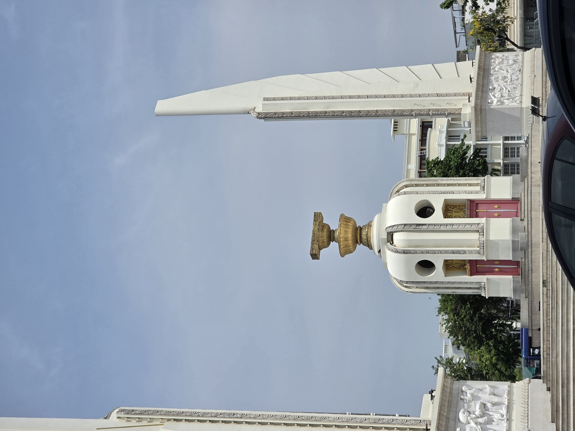 The image features the Democracy Monument in Bangkok, Thailand, captured on a clear day with a light blue sky. The monument stands prominently, characterized by a large, circular white base structure topped with an elaborate, golden, urn-shaped pedestal. Four imposing, white, wing-like structures extend outwards and upwards from the central base, each adorned with decorative patterns along its edge.
Around the lower section of the main pedestal, there are sections featuring red panels and intricate golden patterns, possibly indicating decorative doors or symbolic entrances. The monument's base is further embellished with detailed bas-relief sculptures depicting numerous human figures in various activities, suggesting historical or civic narratives.
In the background, multi-story buildings with numerous windows and lush green trees are visible on both sides of the monument. A dark-colored vehicle is partially seen on the far right edge of the frame, indicating the monument's roadside location. The clear, bright blue sky suggests a sunny daytime setting. No people are explicitly visible, and no specific event or activity is taking place. There is no discernible text in the image.