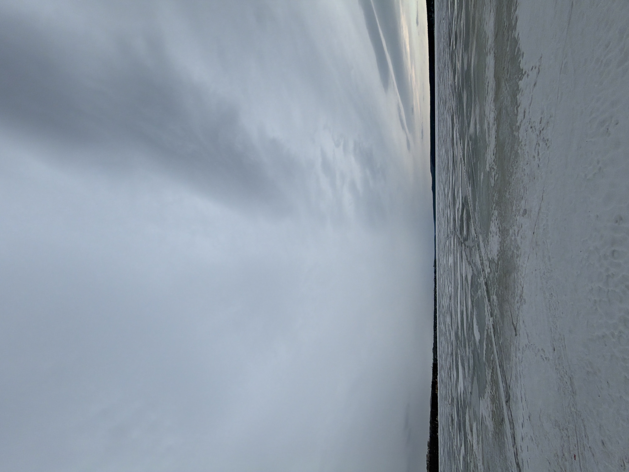 The image shows a scene in Votkinsk, Russia, depicting a frozen body of water, likely a lake or river, covered in snow and ice. The sky is overcast with heavy, grey clouds. A dark line of trees or the shoreline is visible separating the frozen water from the sky. The surface of the snow and ice shows textures and patterns, possibly from wind or previous movement. No people are visible in the image.