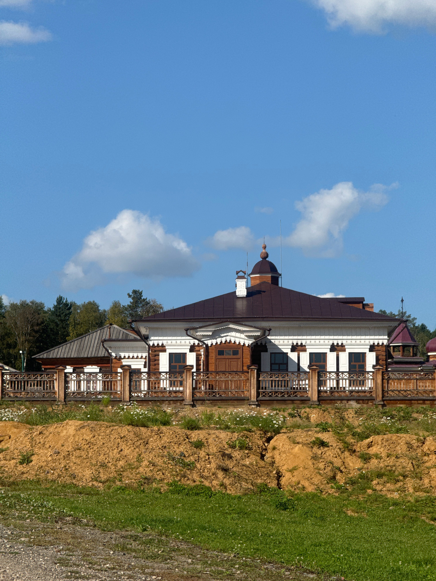The image shows a traditional wooden house with a dark brown roof and white trim, typical of Russian architecture. The house has multiple windows with dark frames and shutters. A wooden fence with an intricate geometric pattern surrounds the property. In front of the fence, there is a mound of dry, reddish-brown soil with sparse green vegetation and small white flowers. Behind the house, a line of green trees can be seen against a bright blue sky with scattered white clouds. The scene appears to be outdoors on a sunny day. The location is identified as Taltsy (Irkutsk), Russia, which is known for its historical architectural complex. There are no people or animals visible in the image.