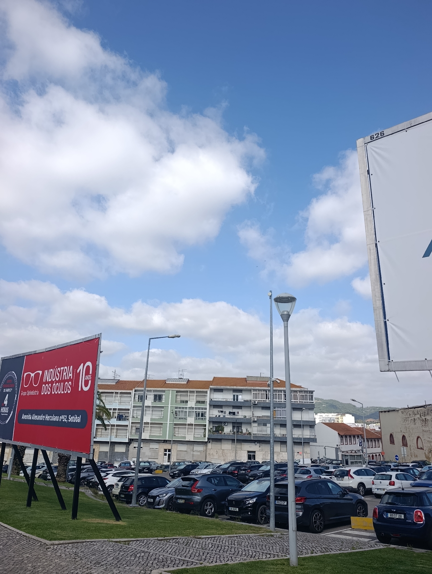 The image depicts a parking lot in Setúbal, Portugal, with a prominent billboard in the foreground advertising "Indústria Dos Óculos" (Eyewear Industry) and "10 Anos" (10 Years). The billboard also states "Avenida Alexandre Herculano n°52, Setúbal", confirming the location. A large white banner with the number "626" is visible on the right side of the image. Numerous cars are parked in the lot. In the background, a multi-story building with balconies and orange-tiled roofs is visible, along with distant hills. The sky is blue with scattered white clouds. There are no people visible in the image.