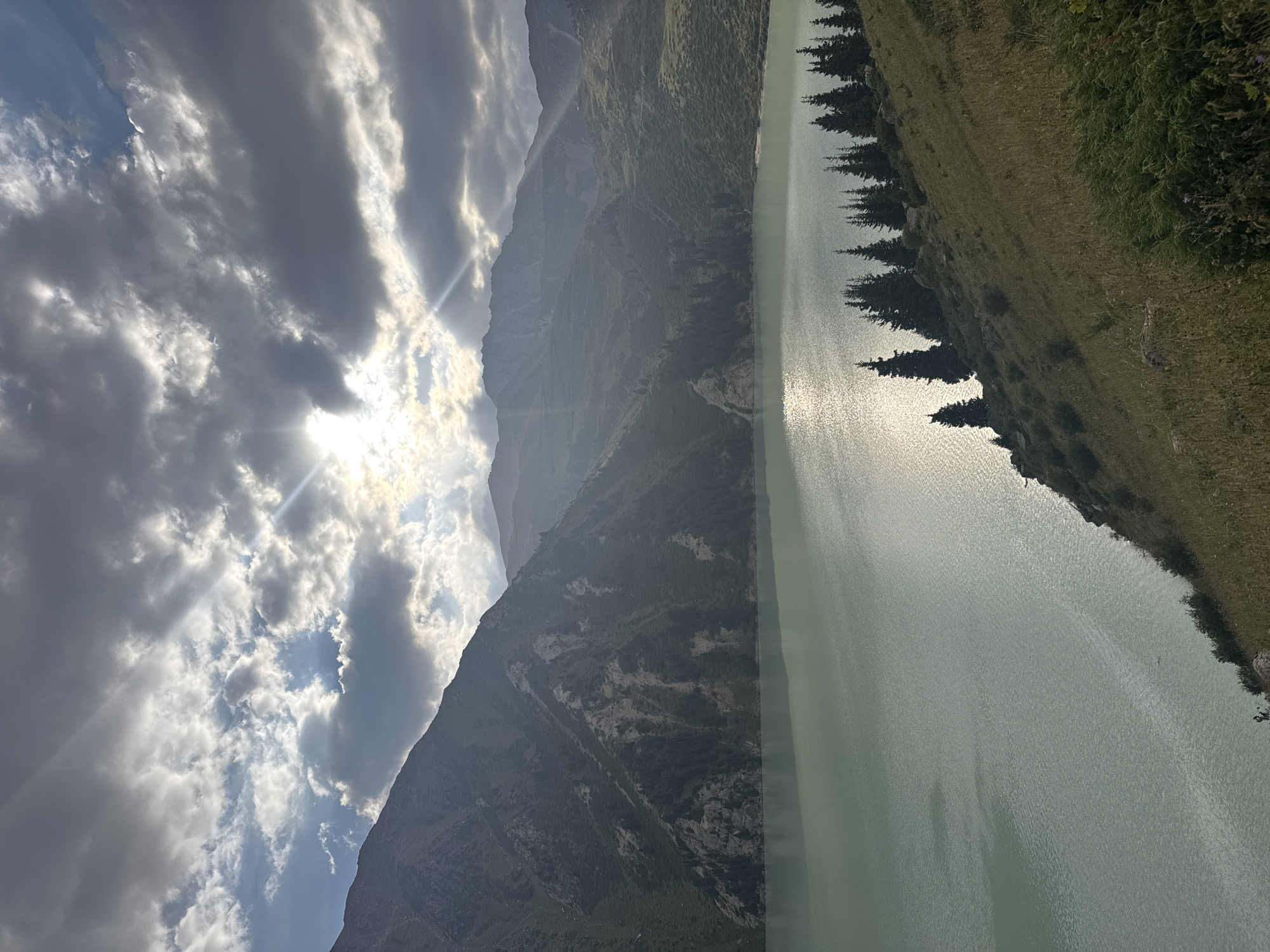The image depicts a panoramic view of Big Almaty Lake, a stunning natural reservoir situated in the Trans-Ili Alatau mountains, approximately 15 kilometers south of Almaty, Kazakhstan. The lake's tranquil, turquoise waters dominate the foreground, reflecting the dramatic interplay of sunlight and clouds in the sky. Jagged, imposing mountains rise steeply from the lake's edge, their slopes carpeted with dense evergreen forests and patches of rocky terrain. The sky above is a canvas of dynamic cloud formations, with sunbeams piercing through gaps, creating an ethereal glow that illuminates the mountainous landscape. The overall impression is one of serene, majestic natural beauty, characterized by the stark contrast between the calm water, rugged mountains, and the dramatic sky. There are no people or animals visible in the scene.
