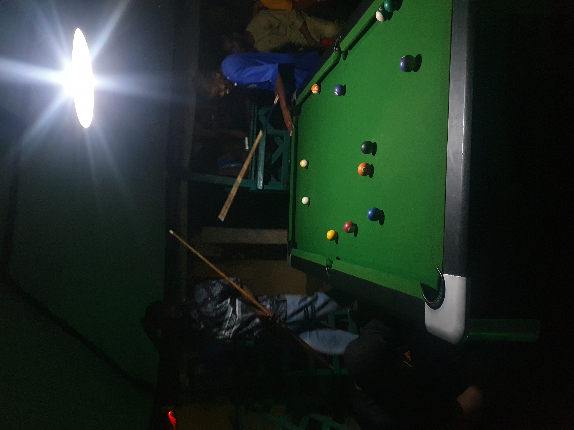 The image depicts a dimly lit indoor scene, likely a bar or recreational area, with a prominent pool table. A bright, oval light fixture on the left casts a strong glare. Several people are present, though their faces are largely obscured by the low light and the angle of the photograph. One individual in a blue shirt is actively playing pool, positioned behind the table with a cue stick in hand. The pool table is green, with various colored balls scattered across its surface, including solid colors and striped balls, along with a black "8 ball" and some red and yellow balls.
Another person, wearing a patterned shirt and jeans, is lying down or sitting on a bench or structure near the pool table, with a pool cue resting beside them. In the background, other individuals are visible, some seated and others standing, further suggesting a social gathering. The overall atmosphere appears casual and relaxed, indicative of a leisure activity.
The location is Lokoja, Nigeria. The time of day is indeterminate due to the artificial lighting and darkness, but the setting suggests an evening or nighttime activity. There are no discernible weather cues. The image lacks any visible text.