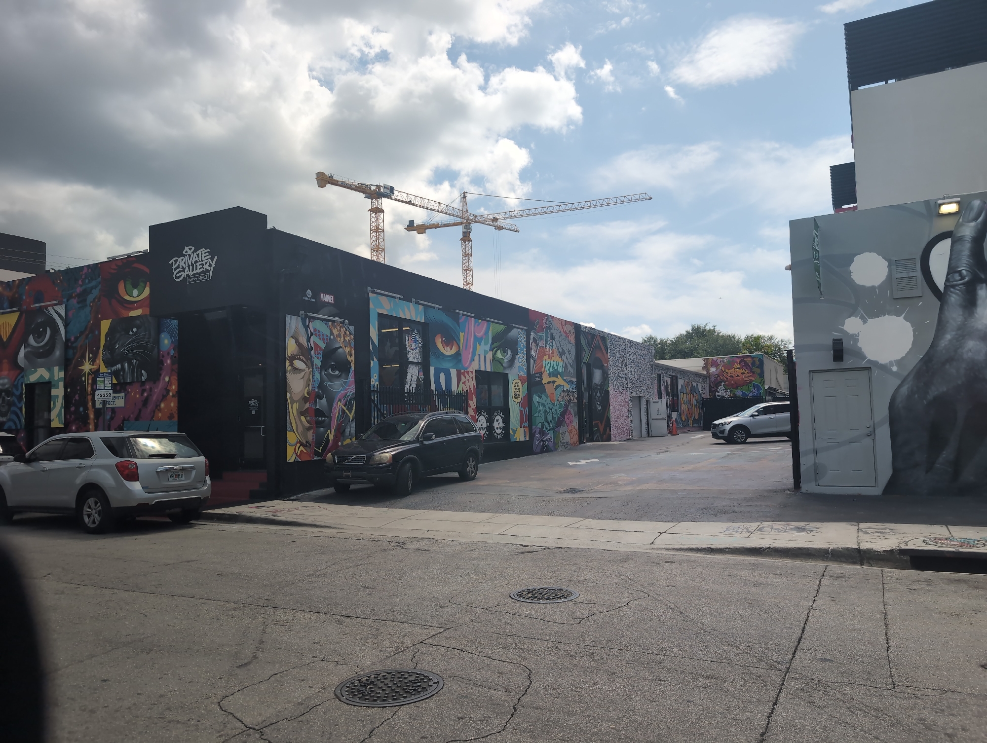 The image captures a street scene in Miami, United States of America, on a partly cloudy day. The primary subjects are two vehicles parked on a cracked asphalt road in front of a building adorned with vibrant graffiti art. One is a silver SUV parked on the left, and a dark-colored SUV is parked on the right, closer to the building. The building itself is black and features a prominent sign that reads "PRIVATE GALLERY" with "ART by ARNO" below it, alongside a "MARVEL" logo. The walls of the building are covered in a mural featuring various artworks, including large eyes and a black panther.
In the background, two large yellow construction cranes are visible against the sky, suggesting ongoing development in the area. Further down the street, more graffiti-covered walls and a white door are visible on another building to the right. The presence of the graffiti, the architecture, and the palm trees visible in the distance all contribute to a distinctly urban, possibly artistic or industrial, district setting.
The lighting suggests it is daytime, with sunlight reflecting off the silver SUV. The clouds indicate dynamic weather conditions. No people are directly visible in the image, but the presence of the gallery and the cars implies human activity in the vicinity. The overall mood of the image is energetic due to the colorful street art, contrasted with the industrial feel of the construction cranes.
