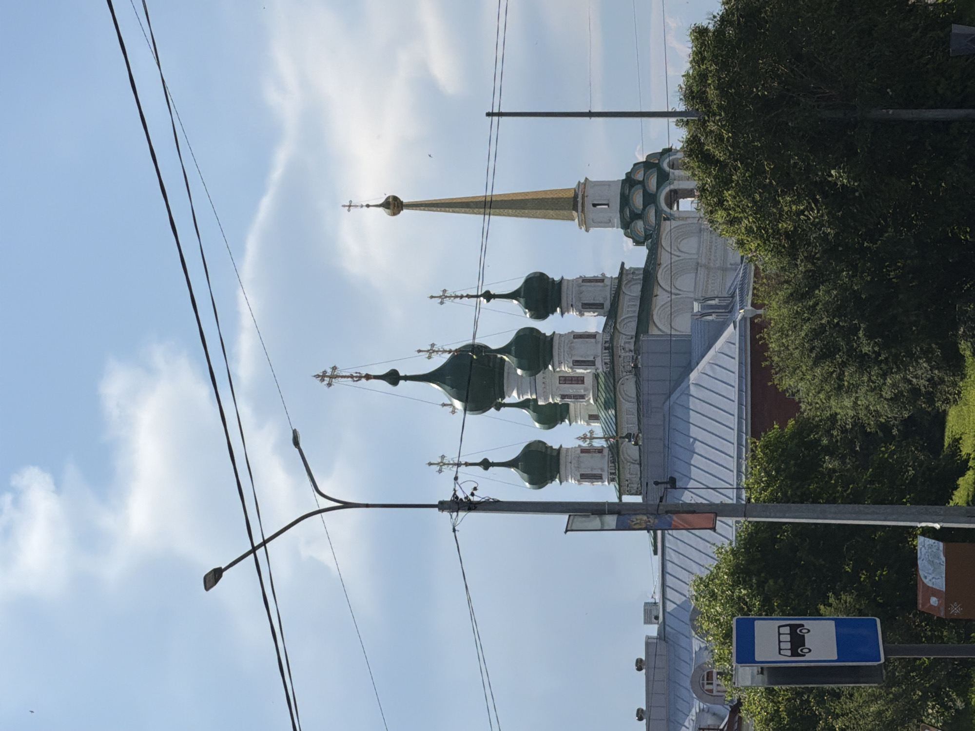 The image captures a view of the Holy Trinity Cathedral in Solikamsk, Russia. The cathedral features multiple green onion domes and a tall, golden spire, characteristic of Russian Orthodox architecture. In the foreground, there is a bus stop sign, a street lamp, and some overhead power lines, suggesting an urban setting. Lush green trees partially obscure the lower portion of the cathedral and surrounding buildings. The sky is partly cloudy, with a few birds visible. The overall scene depicts a sunny day with a mix of architectural and natural elements.