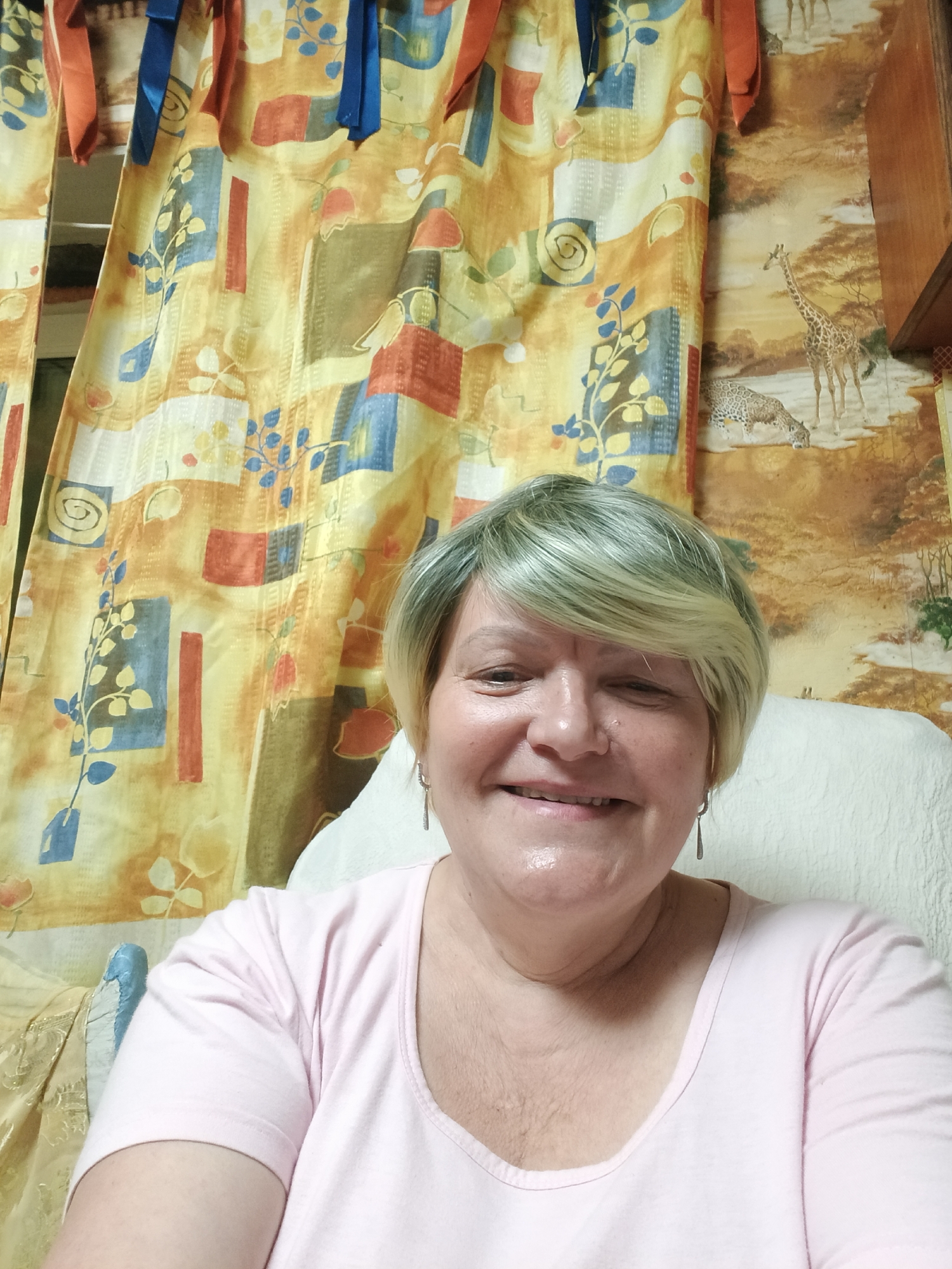 A woman is posing for a selfie. She is wearing a light pink t-shirt and has short blonde hair. The background includes a patterned curtain and a wall covering with a giraffe design. The location is Красный Маяк, Russia.