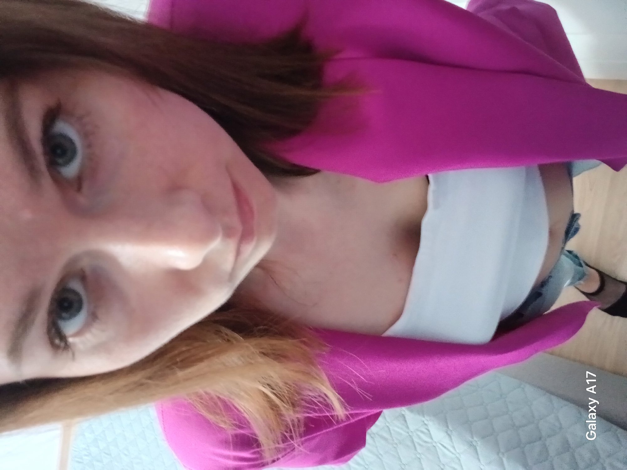 A woman with light skin, light eyes, and brown and blonde hair is facing the camera. She wears a bright pink blazer, a white top, and blue jeans. Black hosiery is visible on one leg. The photo was taken indoors, with a bed visible in the background. The location appears to be Yekaterinburg, Russia, as indicated by the camera name "Galaxy A17" in the lower right corner.