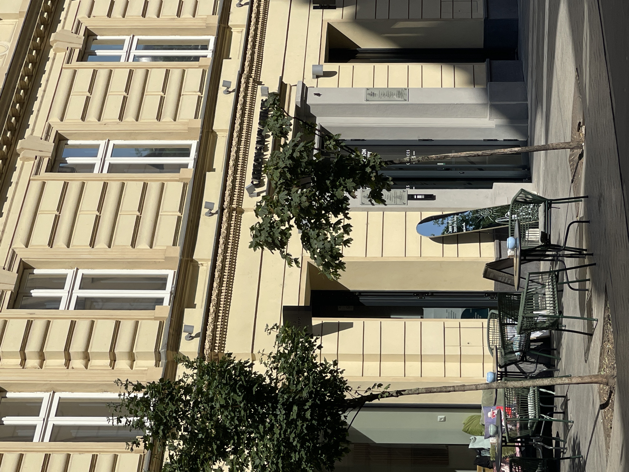 The image captures an outdoor seating area of a cafe located in Bratislava, Slovakia. The cafe is situated on the ground floor of a light yellow, multi-story building with ornate architectural detailing, including layered facade elements and decorative moldings above the windows. Several trees with green foliage are strategically placed around the seating area, providing shade.
The seating area consists of dark green metal tables and chairs. One table is set with blue bowls, and another has a dark-colored table runner. Cushions, some green and one with a patterned design, are visible on a bench or sofa in the background, suggesting the cafe is furnished for comfort. A slender tree trunk is prominently positioned in the foreground, with its shadow falling across the paved ground.
Various signs are visible. One is an A-frame sign placed on the pavement, and a tall, oval-shaped mirror leaning against a column displays text including "Summer is here. Season is a happy...". Above the entrance to the building, a dark sign reads "CARLTON SAVOY OFFICE". Two framed notices are mounted on a pillar near the entrance. The overall scene conveys a relaxed atmosphere on a sunny day, with ample natural light casting distinct shadows.