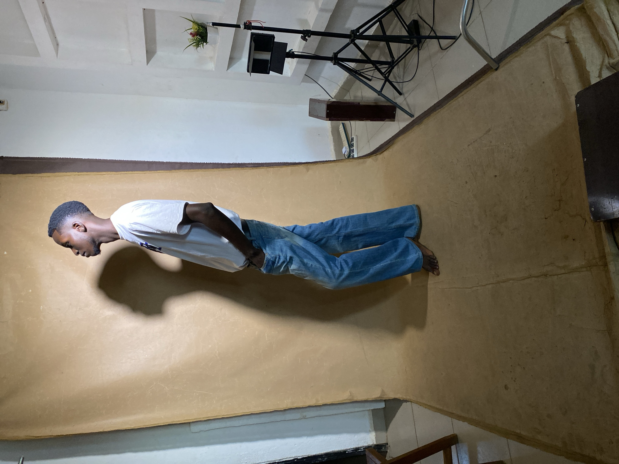 A young Black man is depicted posing in a simple indoor photo studio setup in Bukuru, Nigeria. He is standing on a light brown or tan seamless paper or fabric backdrop that curves from the floor up the wall. The man wears a white t-shirt and blue jeans, with his bare feet visible. He is positioned facing left, looking downwards with his head slightly bowed, and his left hand is in his pocket.
A prominent, dark shadow of the man is cast on the brown backdrop behind him, indicating a primary light source in front of him. To the right of the man, black photography or lighting equipment, including a tripod stand with a rectangular light panel and visible wires, is set up. The underlying floor, visible where the backdrop ends, is tiled. The walls and ceiling are white, with the ceiling featuring recessed rectangular panels. Decorative elements include a small white pot with green and red artificial flowers hanging from the ceiling and a dark brown rectangular box mounted on the wall. Part of a dark wooden object is also visible in the bottom right corner. The man's expression is neutral. No visible text is present in the image.
