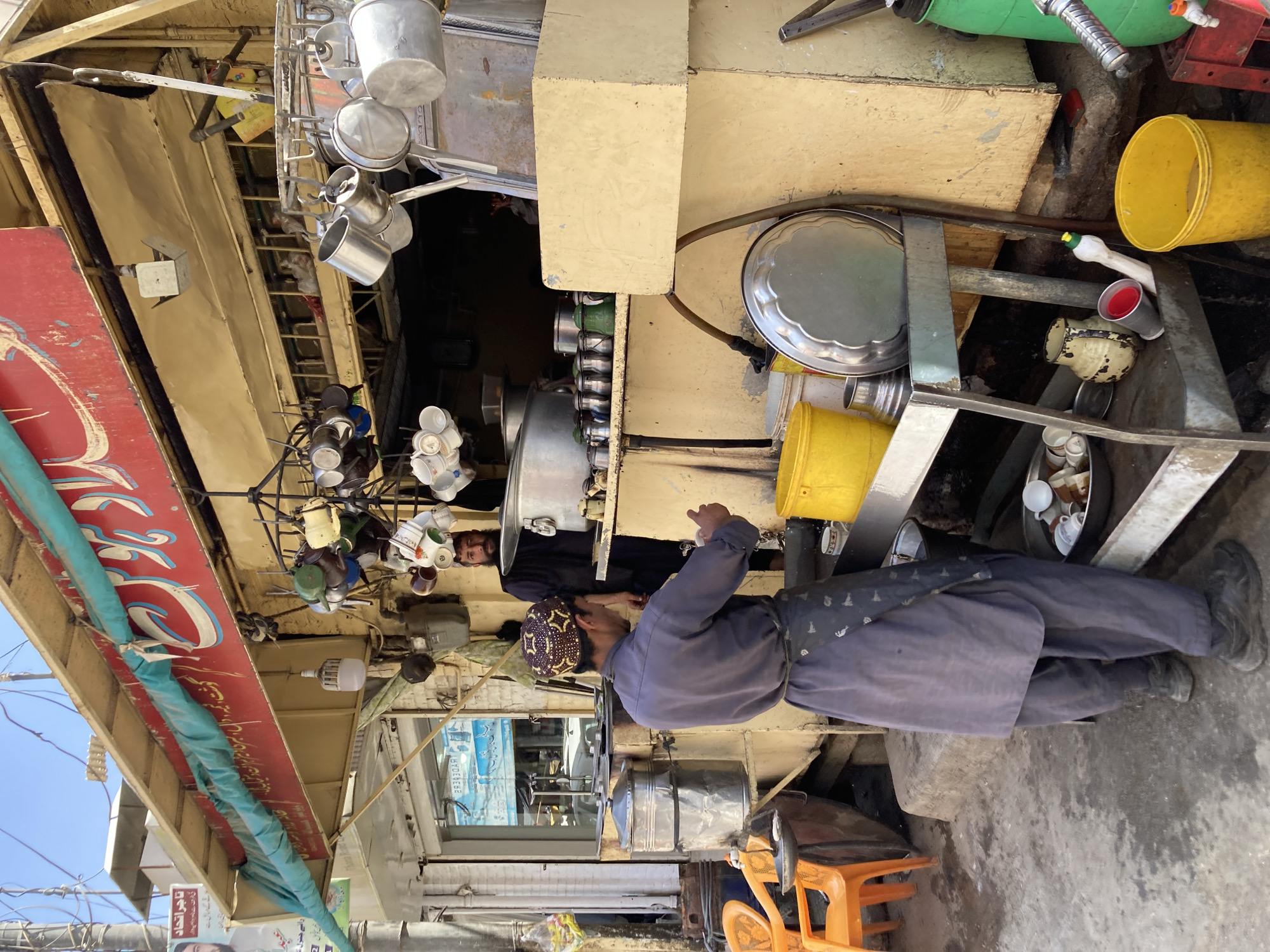 A man wearing a traditional hat and blue outfit stands next to a street vendor stall in Quetta City Tehsil, Pakistan. The stall is cluttered with cooking pots, buckets, and other items. The man appears to be interacting with the stall or another person partially visible behind the stall. The surrounding area is a busy street with shops and signage.