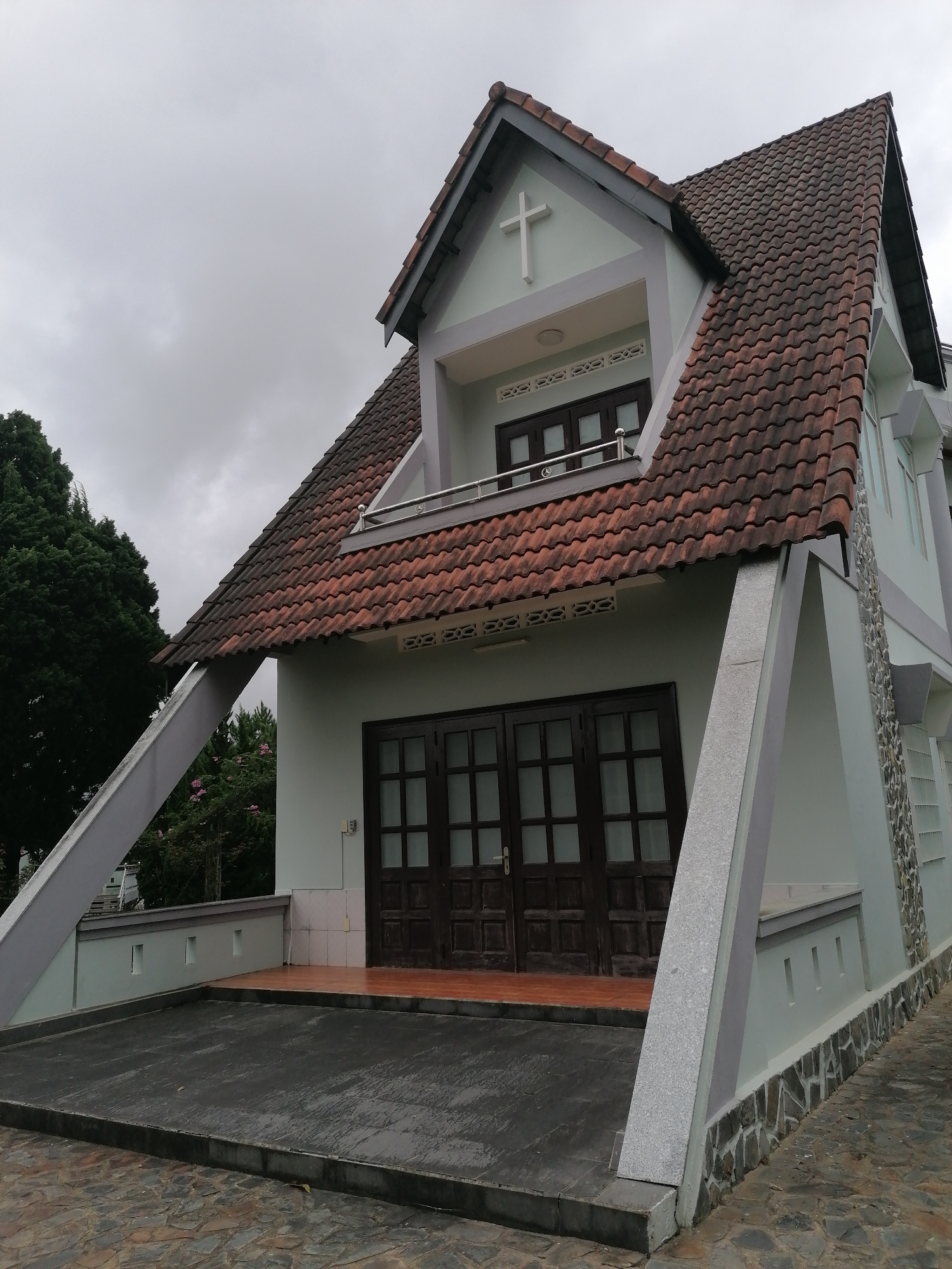 The image shows a light green building with a reddish-brown tiled roof, likely a church, in Lạc Dương, Vietnam. The building features a steep, multi-gabled roof with visible eaves. A white cross is mounted on the upper front gable. The building has large, dark-wood double doors with glass panes, and several windows are visible. Angular, light-colored beams support the roof structure, extending outwards from the building. The ground in front of the building is paved with dark-colored concrete and stone tiles. A large, dark green tree is visible to the left of the building. The sky is overcast and grey.