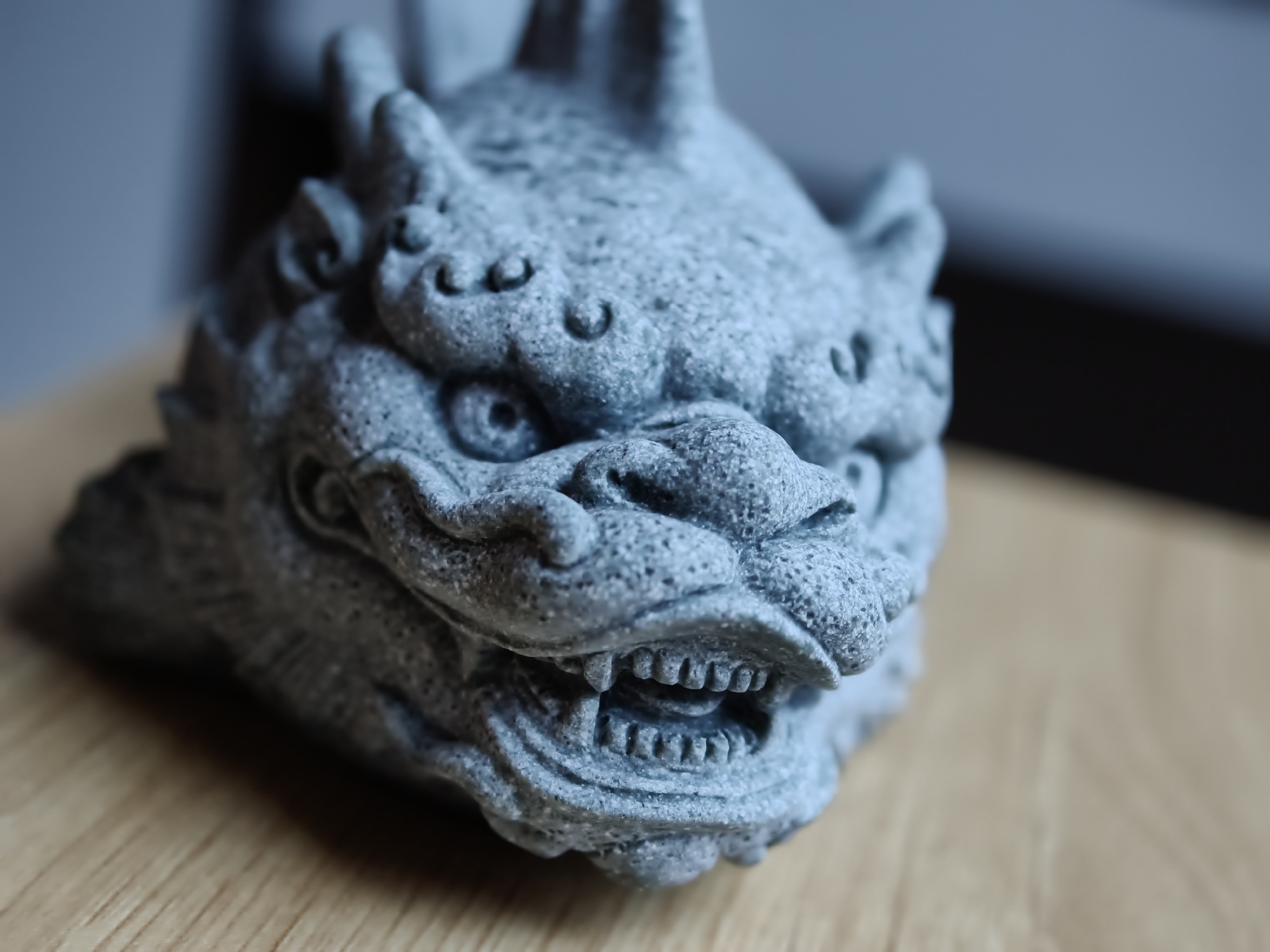 A close-up, shallow depth-of-field photograph captures a detailed, textured, gray stone sculpture of a mythical creature's head. The sculpture, likely a dragon or lion, features intricate detailing such as scaly skin, prominent teeth bared in a fierce expression, and curled embellishments around the eyes and head. The object is positioned on a light brown wooden surface, which appears to be a table or countertop. The background is blurred, with a darker, out-of-focus area to the right and a lighter, muted blue area to the left. The lighting casts subtle shadows, highlighting the texture of the sculpture. There are no people, animals, or visible signs of location specific to Đà Nẵng, Vietnam.