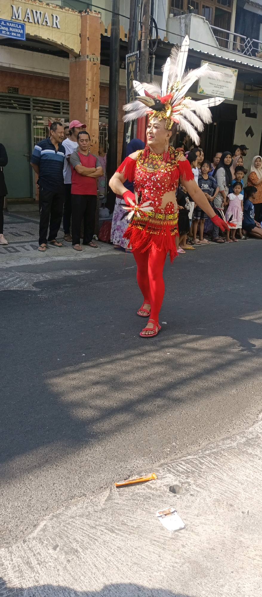 A street celebration is taking place in Malang, Indonesia, with a person in a vibrant red costume and a feathered headdress performing in the foreground. The performer, wearing red leggings and sandals, is adorned with intricate beading and fringe on their top and skirt. Spectators, including men, women, and children, are gathered on the sidewalk and street, observing the event. Buildings with signage, including one that reads "JL. NAKULA GANG. MAWAR RT. 01 RW. 05", line the street. The ground is paved asphalt, with shadows cast from overhead structures. Some litter, including a cigarette pack and what appears to be a wrapper, is visible on the street.