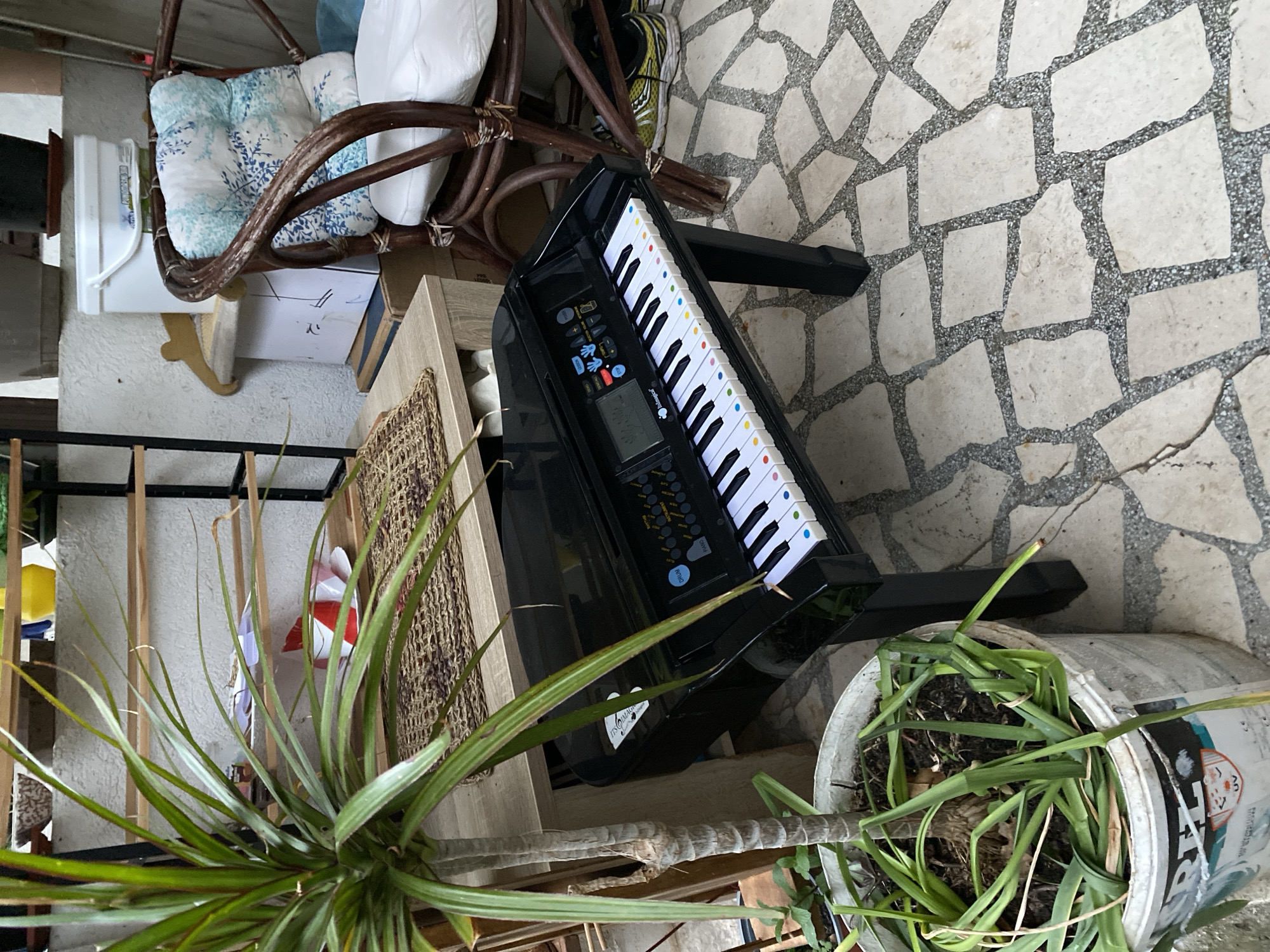 The image shows a cluttered outdoor scene, possibly a patio or terrace. A black toy keyboard with colored keys is positioned on a light-colored table or surface. A potted plant with long, spiky leaves sits nearby, and another potted plant with broad green leaves is visible on the right. In the background, there is a rattan chair with patterned cushions. The ground is paved with irregular, light-colored stone tiles. The location is plausibly in Budva, Montenegro, given the common architectural styles of outdoor spaces.