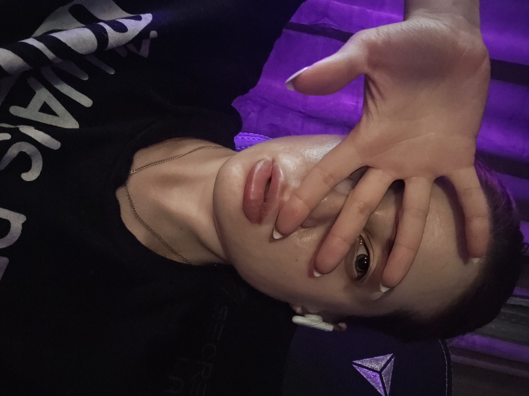 A close-up, rotated view of a person with fair skin, covering one eye with their hand. The person is wearing a black t-shirt with white lettering and a thin gold necklace. Their fingernails are painted with white tips. There is a purple curtain in the background and part of a chair with a logo in the lower right corner of the image. The location is not discernible as Burgas, Bulgaria.