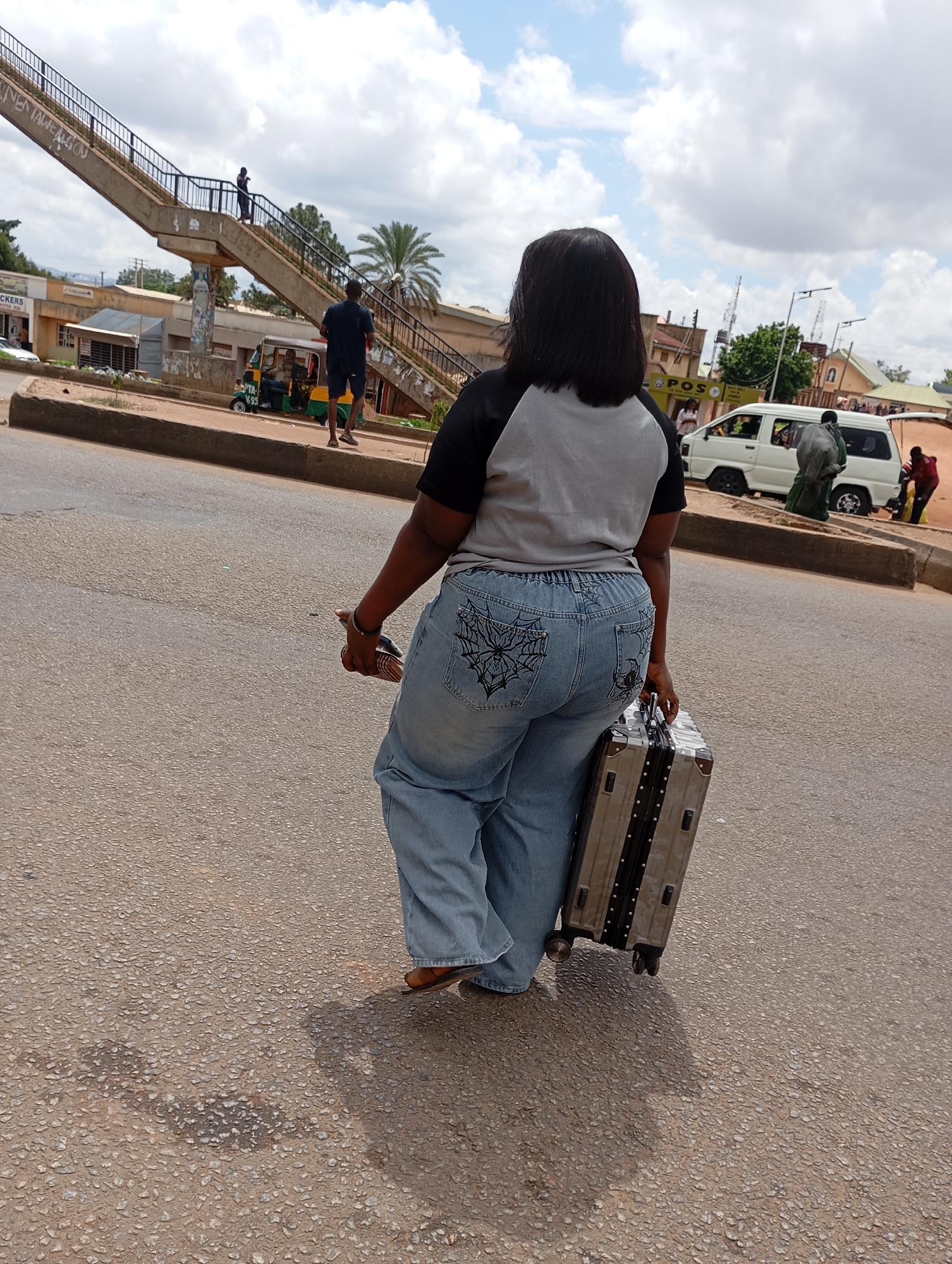 A full-shot, rear-facing medium shot captures a Black woman walking on a paved road in Gura Top, Nigeria. She is wearing a grey and black t-shirt and light blue denim jeans adorned with a spiderweb design on the back pockets. She is holding a brown object in her right hand and pulling a silver suitcase on wheels with her left hand. The road is speckled with small stones. In the background, a concrete pedestrian overpass stretches across the road, with graffiti visible on its side. Several buildings line the roadside, and a white van is parked near one of them. A palm tree stands behind the overpass. The sky is cloudy.