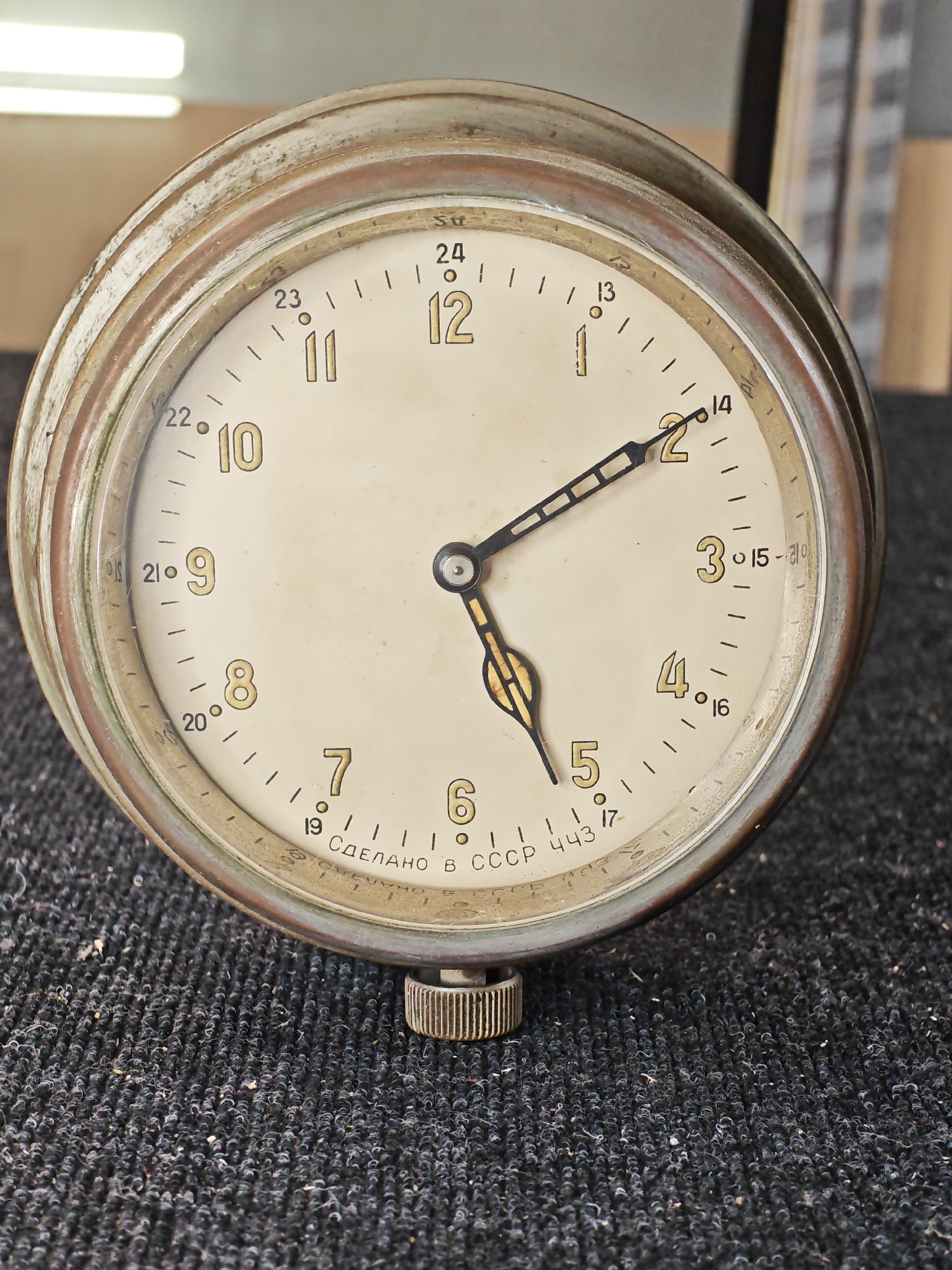 A close-up, eye-level shot shows a vintage Soviet-era clock with a cream-colored face, gold Arabic numerals, and black hands. The clock has a worn, metallic bezel and is set on a dark, textured surface. The time displayed on the clock is approximately 1:59:30. The phrase "СДЕЛАНО В СССР 443" is visible on the lower part of the clock face, indicating it was made in the USSR. The background is out of focus, showing a blurry interior with hints of lighting fixtures and shelving. The specific location within Ждановіцкі сельскі Савет, Belarus, is not discernible from the visual information provided in the image.