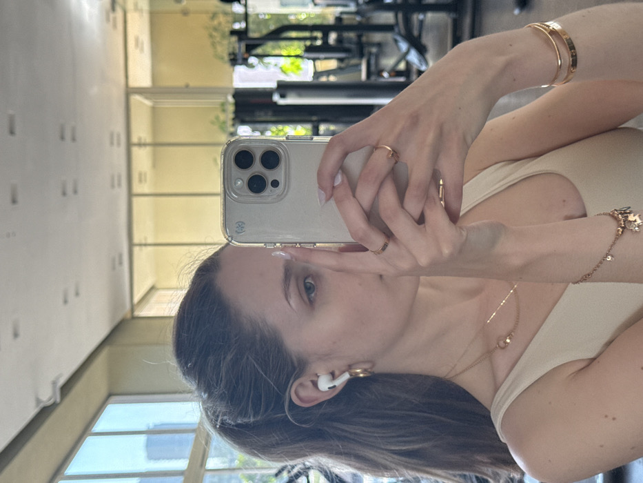 A female subject is depicted in a mirror selfie taken inside a modern gym, obscuring part of her face with a silver-colored smartphone held in both hands. She has light skin, brown hair tied in a high ponytail, and wears a light-colored, sleeveless top. Visible accessories include white wireless earbuds and gold hoop earrings in her left ear, a delicate gold necklace, multiple gold rings on her left hand, a gold charm bracelet on her left wrist, and two gold bangle bracelets on her right wrist. Her fingernails are manicured with a light polish. The smartphone, likely an iPhone model with a triple camera array, is encased in a clear protective cover. The background, seen in reflection, reveals a bright gym interior with large windows to the left, through which urban buildings and green foliage are discernible. Various dark-colored gym equipment is partially visible and out of focus in the mirrored space. The setting is consistent with a contemporary fitness center located within an urban environment in Mexico City; however, the precise street address or specific neighborhood within the city cannot be determined from the visual information provided.