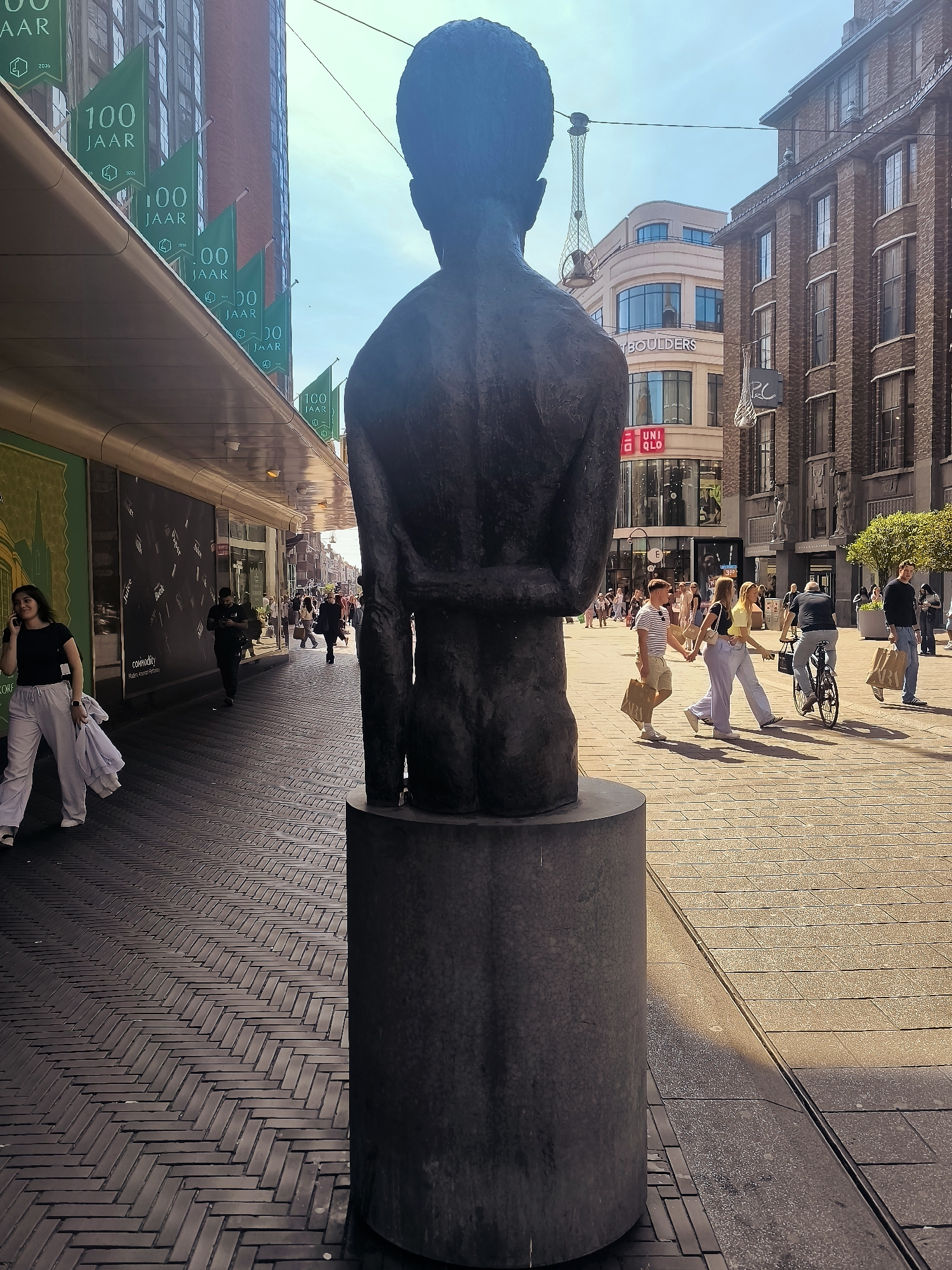 The image shows a street scene in the city of The Hague, Netherlands, likely on a shopping street given the presence of stores like Uniqlo and "Boulders." In the foreground, a dark bronze statue of a person's torso and head is prominently displayed on a cylindrical pedestal. The statue is viewed from behind. To the left of the statue, a woman in white pants and a black top is walking away from the camera, talking on her phone and carrying white bags. Behind her, a man in a dark uniform appears to be working. Further down the street, several other people are walking, some carrying shopping bags.
The buildings on either side of the street are a mix of architectural styles. The building on the left has a modern awning with green flags bearing the text "100 JAAR" (100 years) and the year "2026" visible. The building on the right is made of dark brick with numerous windows.
The street is paved with light-colored bricks laid in a herringbone pattern. Sunlight casts shadows across the pavement, indicating it is daytime. The sky is clear and blue. The overall atmosphere appears to be that of a busy urban shopping district on a pleasant day.
