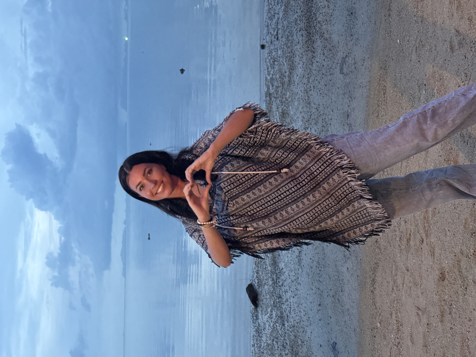A young woman stands on a beach in Baan Sri Thanu, Thailand, making a heart shape with her hands. She has long, dark hair and is wearing light jeans and a poncho-style top with fringe. The beach is sandy and meets the water of a bay under a cloudy, blue sky. The scene is likely taken at dusk.