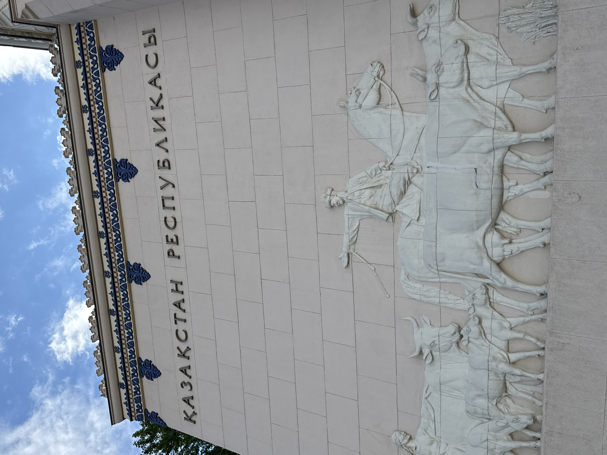 The image shows a relief sculpture on the facade of the Kazakhstan Pavilion at VDNKh in Moscow, Russia. The sculpture depicts a procession of people and livestock, with a man on horseback leading a group of cows and calves. The man on horseback is dressed in traditional Kazakh attire and is holding a whip. A woman is shown to the left of the cows, possibly representing a shepherdess. Above the sculpture, the words "КАЗАҚСТАН РЕСПУБЛИКАСЫ" (Kazakhstan Republic) are inscribed in Cyrillic letters. The building facade is made of light-colored stone blocks, and decorative blue patterns are visible along the top of the wall. The sky above is partly cloudy.