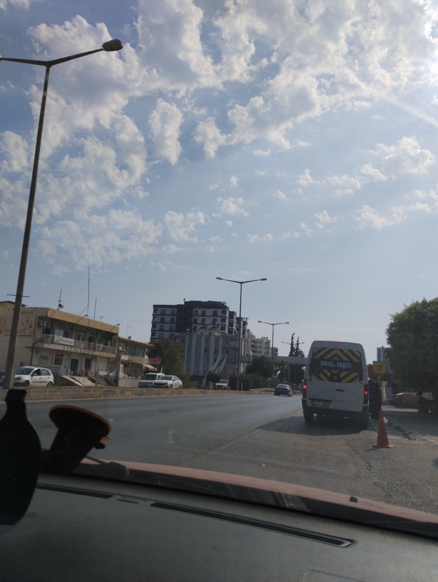 The image captures a street scene in Erdemli, Turkey, under a bright, partially cloudy sky. In the foreground, the dashboard of a vehicle is visible, with a suction cup mount for a device. The road ahead shows moderate traffic. A white van with the text "OKUL TAŞITI" (school transport) written on its rear is a prominent subject, stopped on the right side of the road, possibly indicating a school pickup or drop-off. An individual in dark clothing stands near the van. To the left of the road, buildings line the street, including a beige, two-story structure with businesses on the ground floor and a larger, modern building with dark accents further down. Streetlights are positioned along the road. Several cars are parked or driving on the street, including a white hatchback and other sedans. Trees and vegetation are visible on the right side of the image. The overall impression is a snapshot of everyday urban life in Erdemli.