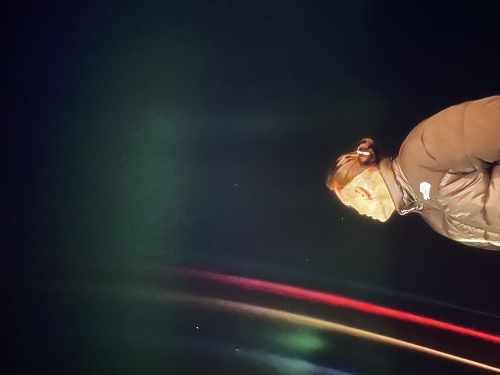 A person wearing a brown puffer jacket and with their hair in a bun is positioned to the right of the image. The background consists of a dark green and black sky with multiple horizontal streaks of red, orange, yellow, green, and blue light. The location is Unknown, Unknown.
