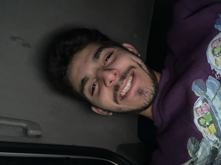 A young man smiles at the camera from the passenger seat of a car. He has dark curly hair, brown eyes, and a trimmed beard. The car interior is dimly lit. He wears a purple sweatshirt with a cartoon design. The roof of the car and a grab handle are visible above his head.