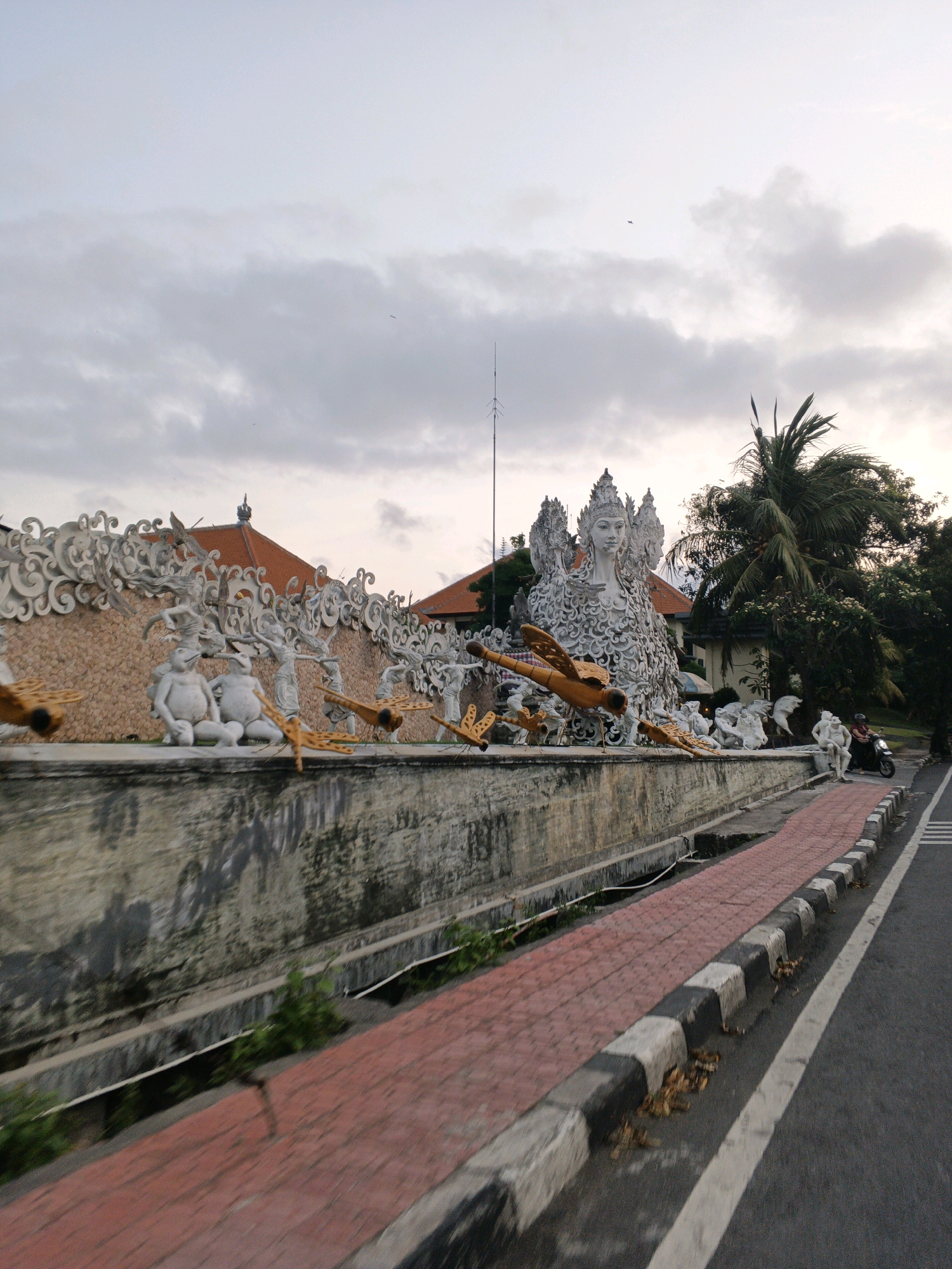 The image depicts a street scene in Denpasar, Indonesia, with a prominent artistic installation on the left side. The installation features a large white sculpture of a woman with elaborate headdress and ornate clothing, surrounded by smaller white figures and stylized yellow dragonfly sculptures. Behind the installation, a textured wall with an orange, pebble-like pattern is visible, topped with decorative white architectural elements. Red-tiled rooftops of buildings are also present. To the right of the installation, a sidewalk made of red bricks runs alongside the road. The road itself is paved and has white lane markings. Trees, including a palm tree, are seen in the background. A person on a black motorcycle is riding on the road, moving away from the camera. The sky is overcast with scattered clouds. The overall impression is a blend of urban infrastructure and cultural artistry.