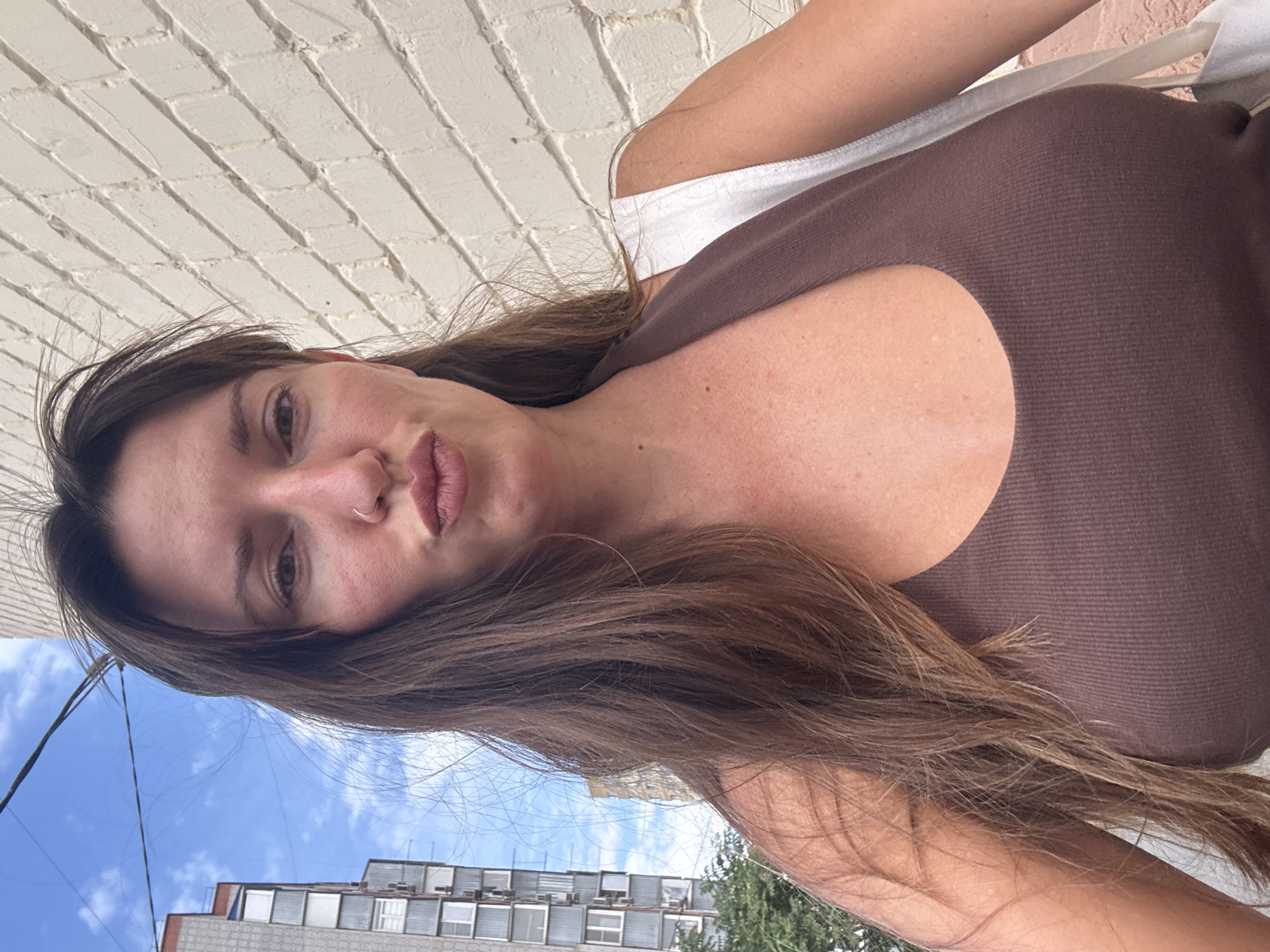 A woman with long brown hair and a nose ring poses for a selfie outdoors in Bataysk, Russia. She is wearing a brown ribbed tank top and a white bag is slung over her shoulder. Behind her, a light-colored brick wall is visible to the right, and a tall apartment building and a blue sky with some clouds are to the left. The overall lighting suggests it is daytime.