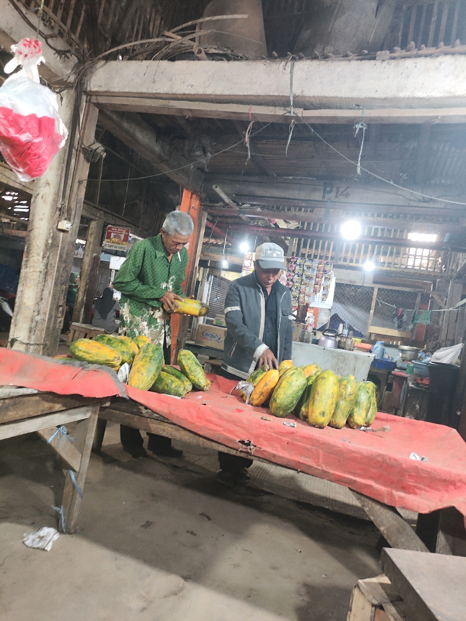 In Karawang, Indonesia, within a dimly lit, open-air market stall constructed from rough wood and concrete, two men are attending to a display of papayas. The stall has a rustic appearance, with exposed beams overhead and a corrugated metal roof partially visible. A red tarp covers a wooden table on which the papayas are neatly arranged. One man, wearing a green patterned shirt and a traditional sarong, holds a papaya and appears to be preparing it, possibly for sale. He has grey hair and a cigarette in his mouth. The other man, dressed in a dark jacket and a light-colored baseball cap, stands behind the table, his hand resting on the fruit. Behind them, shelves are stocked with various packaged goods, and other market activity is suggested by blurred figures in the background. The overall impression is of a busy, local market scene.