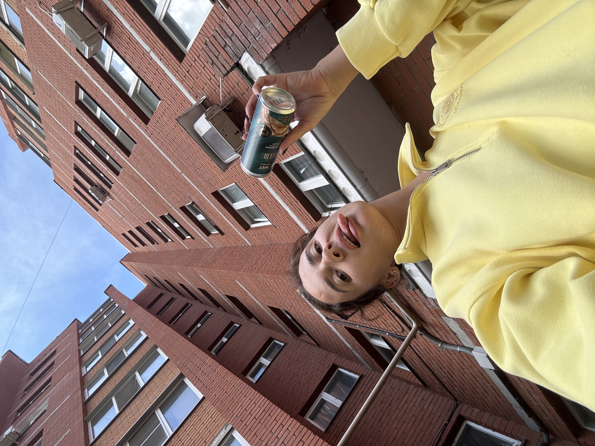A young woman with dark curly hair, wearing a bright yellow quarter-zip sweatshirt, sticks her tongue out at the camera while holding a can of drink in her right hand. She is positioned in the foreground, looking up towards the sky and tall brick buildings. The buildings are characteristic of Soviet-era apartment blocks, with many windows and exterior air conditioning units. The perspective is a low-angle shot, emphasizing the height of the buildings. The sky above is partly cloudy. The scene appears to be an outdoor urban environment, possibly a courtyard or street between apartment buildings in Chaikovsky, Russia, a city known for its characteristic architecture from that period.