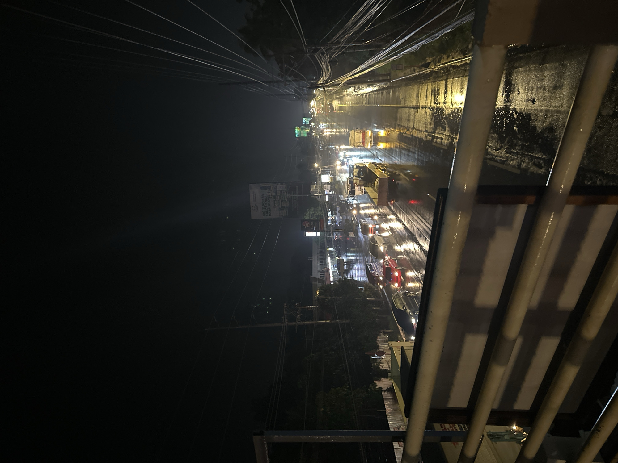 The image depicts a nighttime scene on a rain-slicked road in Davao City, Philippines. The perspective is from an elevated position, likely a pedestrian overpass or bridge, looking down at the traffic. The road is wet and reflective, with the headlights and taillights of vehicles creating streaks of light. Several cars and trucks are visible, some with their hazard lights on. The surrounding environment appears to be a mix of commercial buildings and trees. Utility poles with numerous wires are prominent on the right side of the image, a common sight in urban areas of the Philippines. The overall atmosphere is dark and wet, characteristic of a rainy night.