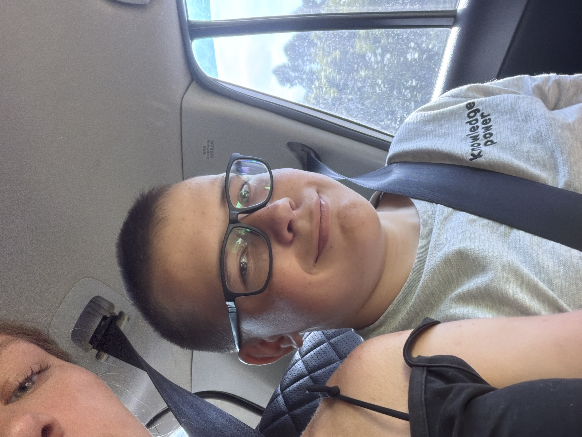 In Kruglovka, Russia, a selfie is taken inside a vehicle. The foreground features a young boy wearing glasses and a gray t-shirt with the words "Knowledge Power" embroidered on it. A seatbelt crosses his chest. To the left of the boy, a portion of another person's face and shoulder are visible, also wearing a black top. The background shows trees and foliage visible through the car window, suggesting an outdoor setting within the city. The interior of the car is also visible, including part of the car's ceiling.