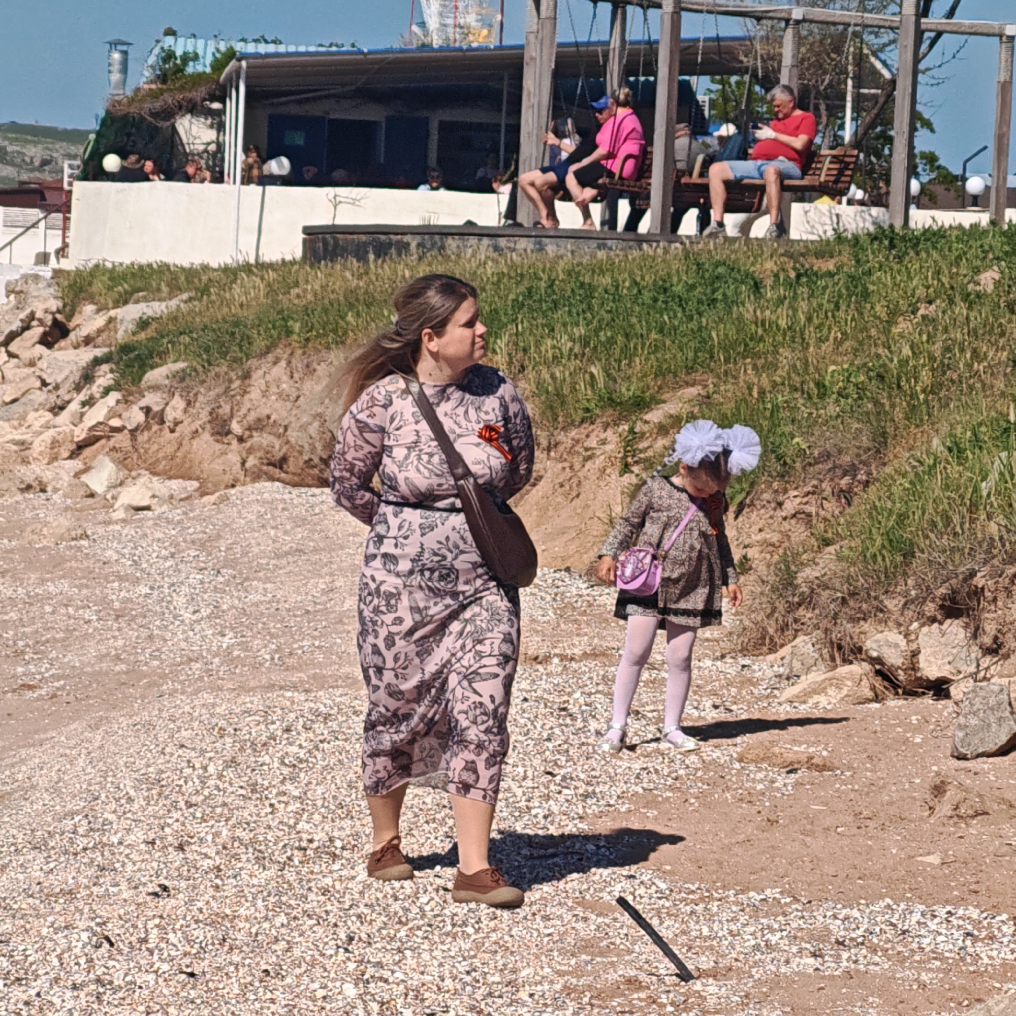 Captured in городское поселение Щелкино, Ukraine, the image shows an outdoor scene on what appears to be a beach or coastal area. A woman, dressed in a floral-patterned dress and brown shoes, is walking with her hands behind her back. She carries a brown shoulder bag. A young girl, dressed in a similar pattern and wearing a large white hair bow, is walking near her, also carrying a small purse. The ground is covered in small rocks and sand. In the background, there is a raised platform with a building structure and individuals seated on benches. A couple is sitting in swings. The overall environment suggests a relaxed, coastal setting on a sunny day.