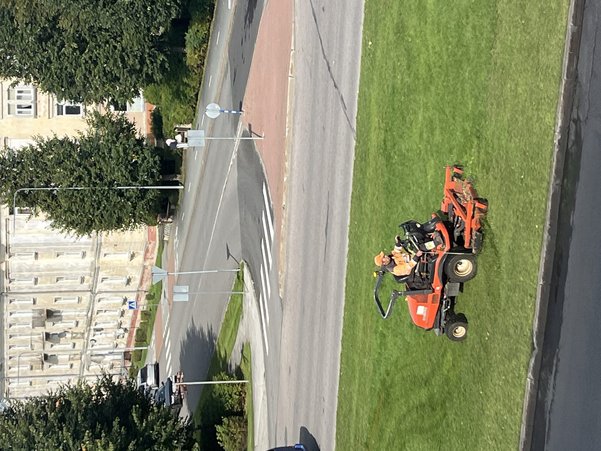 In Kohtla-Järve linn, Estonia, a worker in an orange high-visibility vest and cap operates a red zero-turn lawnmower on a manicured green lawn. The worker is positioned on the right side of the mower, facing forward and holding the steering controls. The lawnmower is actively cutting the grass, with the mower deck engaged and blades rotating. In the background, a street with a pedestrian crossing is visible, with a few cars parked and driving. Beyond the street, a multi-story building with cream-colored walls and numerous windows stands, flanked by mature green trees. The sky is clear and sunny, casting shadows on the ground. The overall scene depicts routine municipal maintenance in an urban setting.