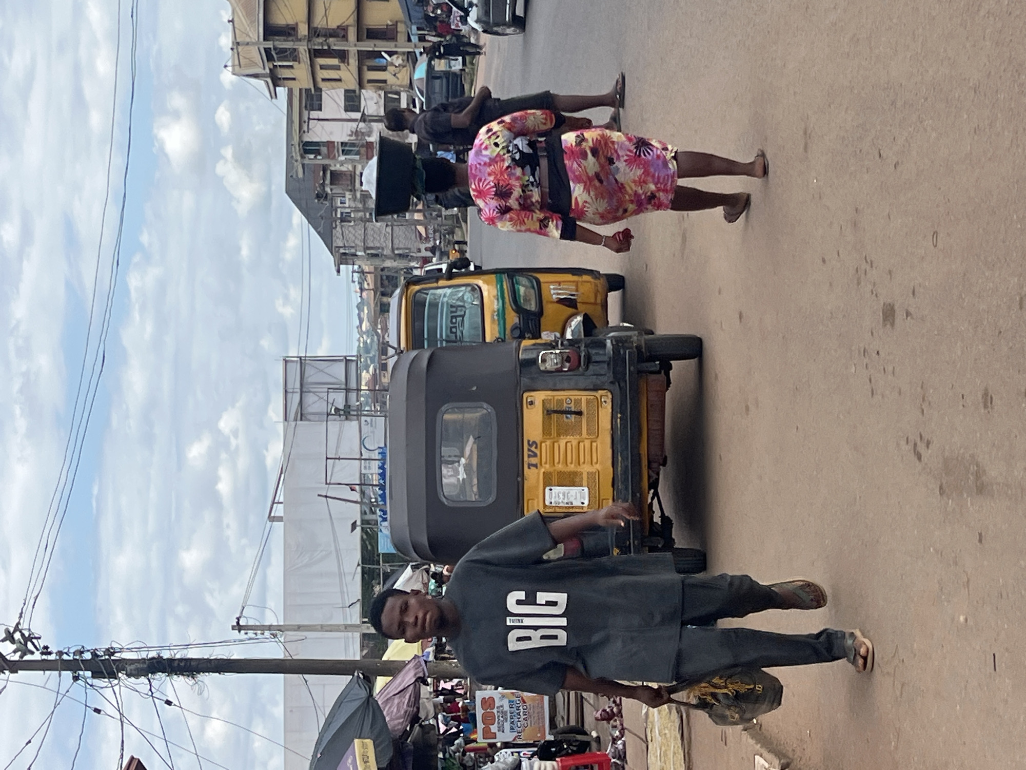 In Nkpor, Nigeria, a busy street scene unfolds under a cloudy sky. A young man wearing a dark t-shirt with "BIG" printed in white walks towards the viewer, carrying a bag in his left hand. He is dressed in dark pants and sandals. Beside him, a yellow auto-rickshaw, a common mode of transport in the region, is parked. To the right, a woman in a colorful floral dress walks away from the camera, balancing a large basin on her head. Another man in a dark t-shirt and shorts walks on the right side of the road. The background shows multi-story buildings, overhead wires, and market stalls, indicating a vibrant urban environment. The road surface is paved, with some scattered debris.