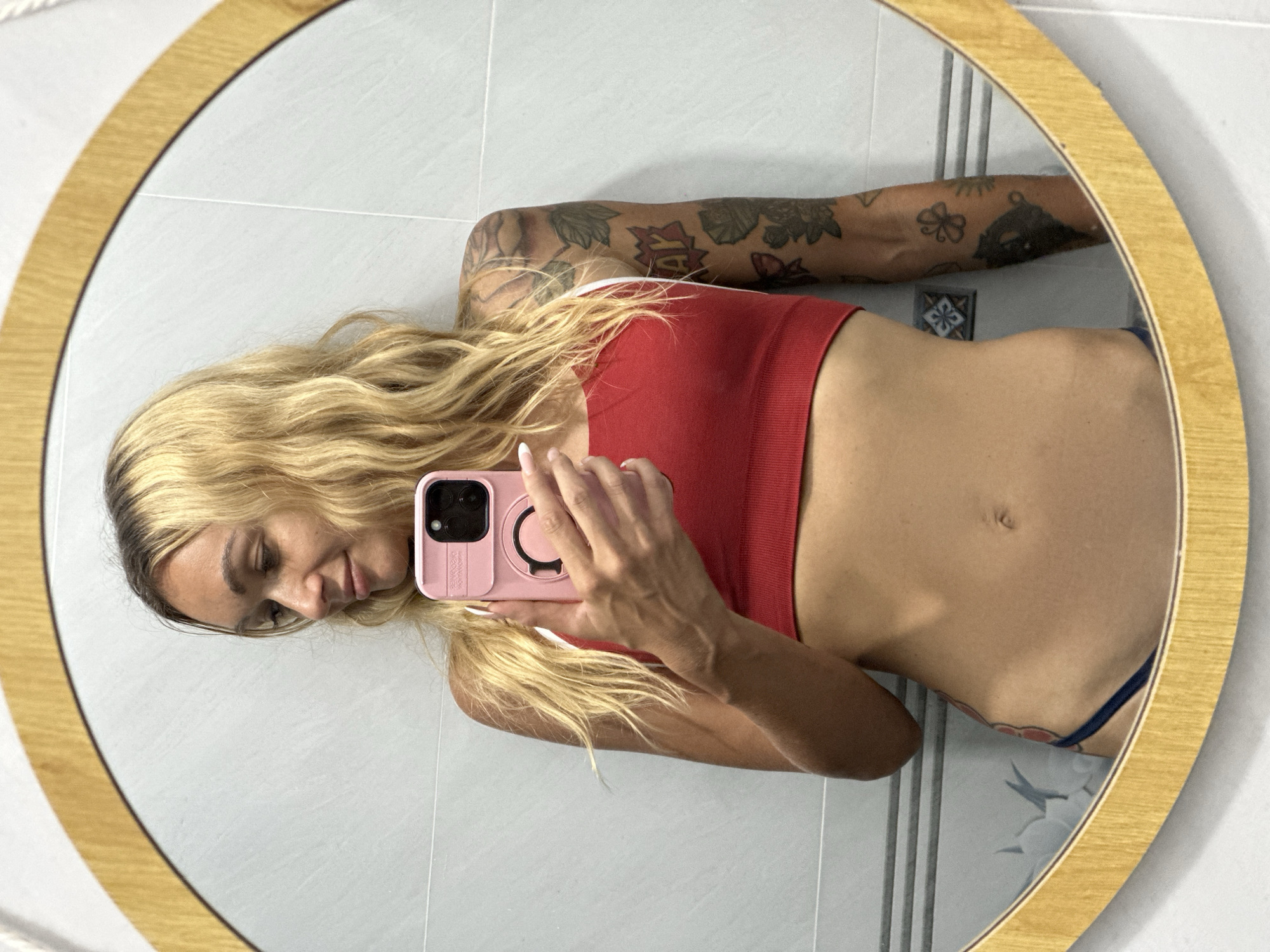 A woman with long, wavy blonde hair is taking a mirror selfie in a bathroom. She is wearing a red crop top and dark blue underwear. She has tattoos on her right arm. The bathroom has white tiled walls and a wooden-framed mirror. The image does not provide enough information to determine its location in Mũi Né, Vietnam.