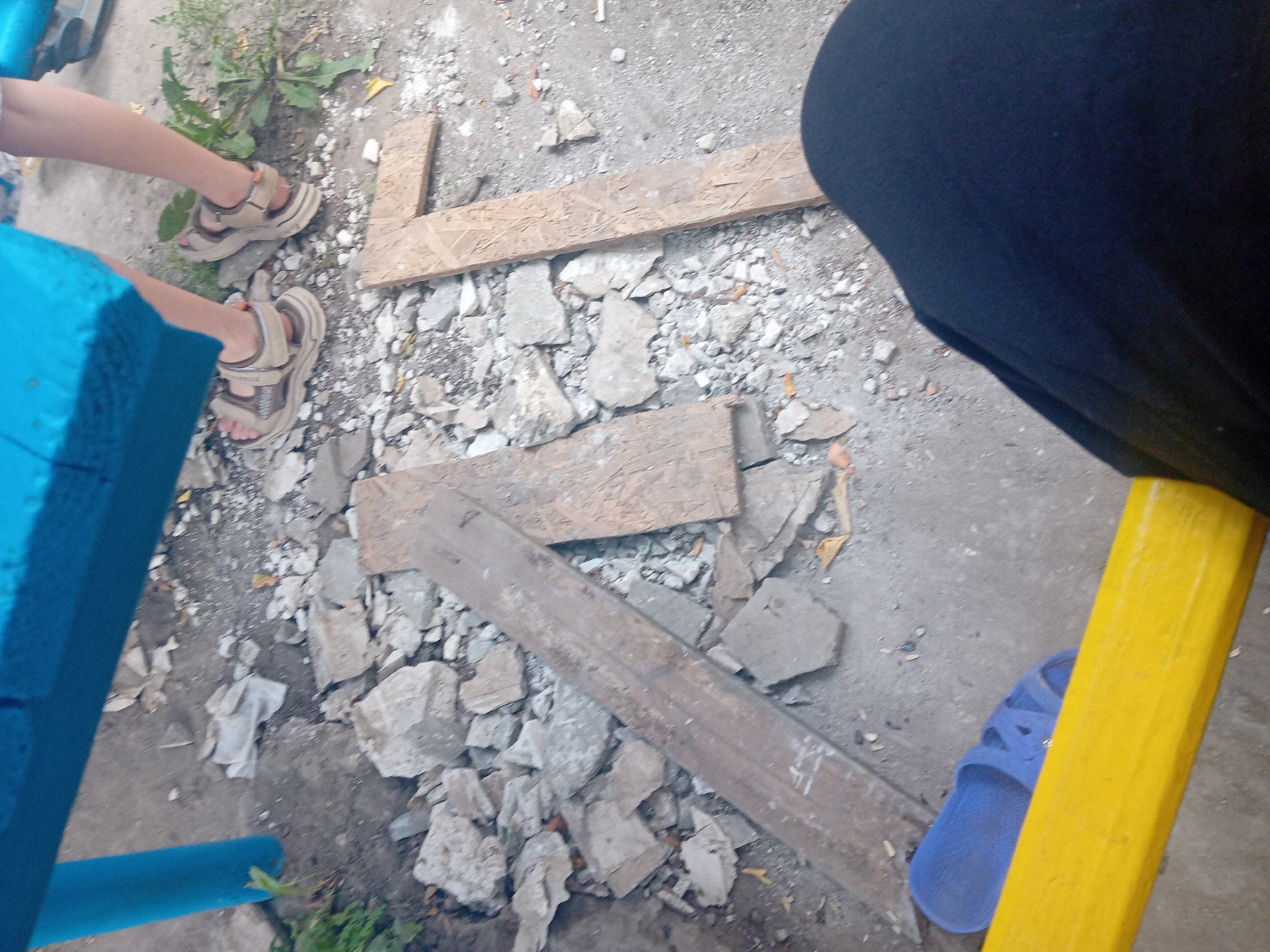 The image depicts an outdoor scene with a ground covered in rubble and debris, suggesting a construction or demolition site. Two individuals are partially visible. One person, wearing tan sandals, stands with their feet positioned over some of the debris. The other person is wearing blue flip-flops and is seated on a yellow object, with their dark-clothed legs in the foreground. The environment includes a blue structure in the upper portion of the image and scattered pieces of wood. The scene appears to be a public space, possibly a playground or a yard, in Chuhuiv, Ukraine, given the context provided. The overall impression is one of disarray and temporary staging.