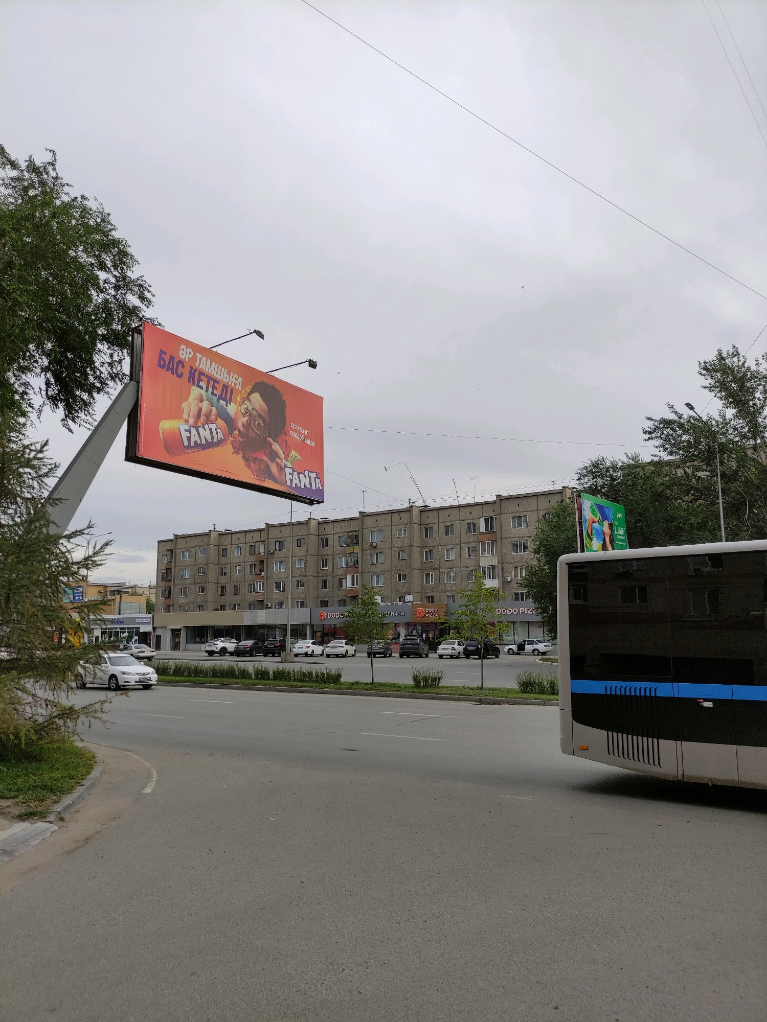 The image shows a wide, grey street in the city of Semey, Kazakhstan. On the right side of the street, a modern bus with tinted windows and a blue stripe is partially visible, moving away from the camera. Further down the street, several cars are parked in front of a building with multiple floors and many windows. On the left side of the street, a large billboard advertising Fanta is prominently displayed. The billboard has an orange background with an image of a person holding a Fanta bottle. Trees are visible on the left side of the image, and the sky is overcast. The overall impression is a typical urban scene in Kazakhstan.