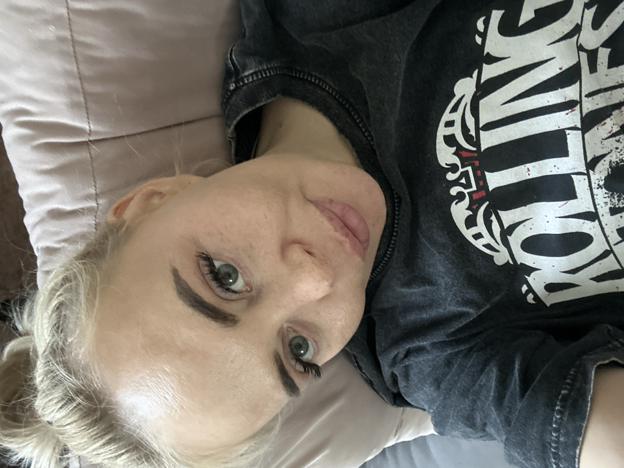 A light-skinned woman with blonde hair and grey-blue eyes is lying down. She appears to be resting on a pillow, and she is wearing a dark grey t-shirt with a white design. The location is Высокий, Russia.