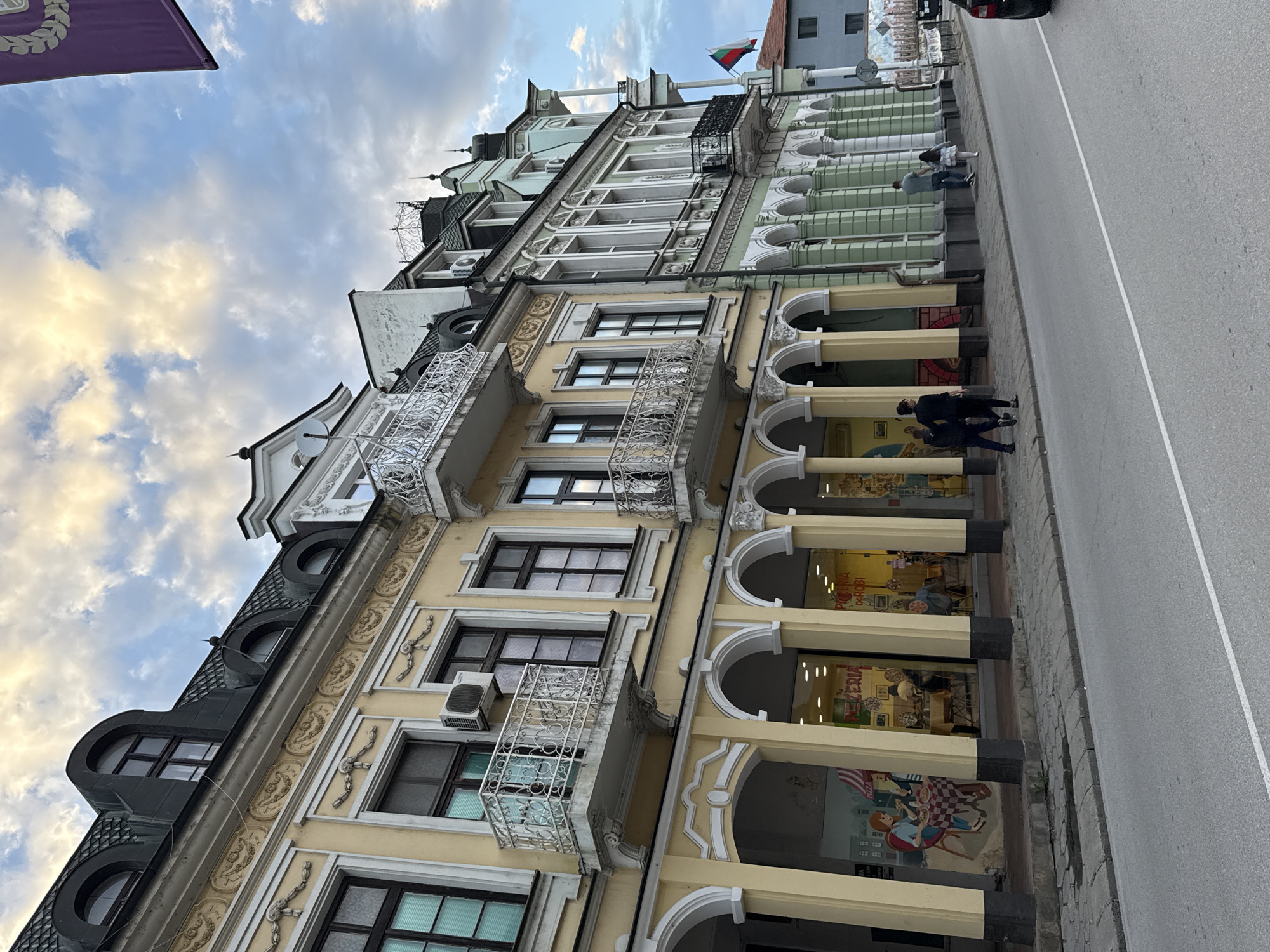 The image shows a street view in the city of Lovech, Bulgaria, featuring ornate, multi-story buildings with a mix of architectural styles. The buildings exhibit features such as arched colonnades at street level, balconies with wrought-iron railings, and detailed ornamentation. The color palette includes yellows, creams, and muted greens. People are walking on the sidewalk next to the buildings. The street itself is made of asphalt and has a visible lane divider. The sky is partly cloudy. A Bulgarian flag is visible atop one of the buildings.