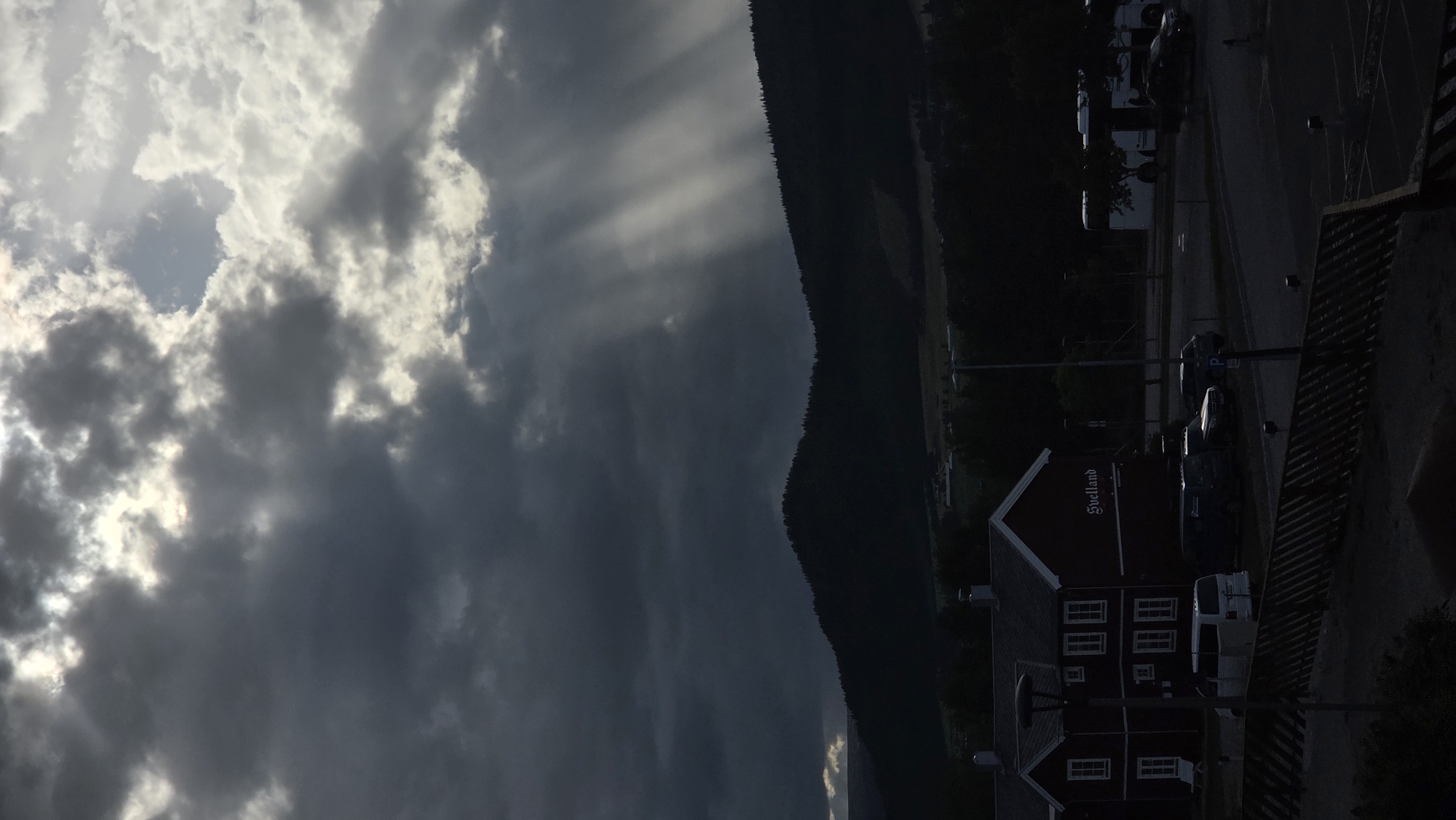 The image depicts a dramatic sky with rays of light breaking through dark clouds, illuminating a silhouetted landscape of hills and trees. In the foreground, a dark red building with white trim, identified by the signage "Svelland", is visible. Several vehicles, including vans and cars, are parked in a lot adjacent to the building. A wooden fence runs across the lower part of the frame. The overall atmosphere is moody and atmospheric, suggesting either the approach of a storm or the aftermath of one. The location is identified as Tynset, Norway.