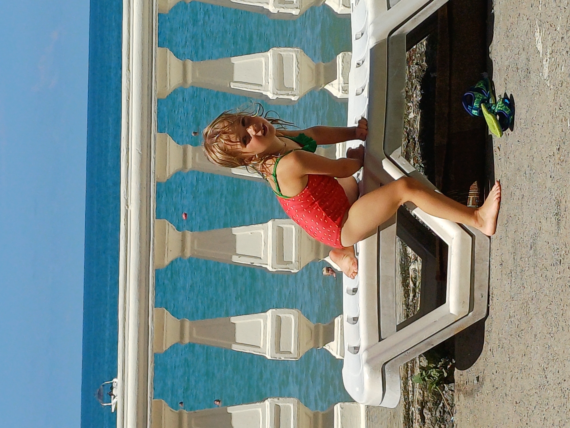 In Sochi, Russia, a young girl with blonde hair, wearing a red swimsuit with a green trim, is positioned on a white plastic lounge chair. She is leaning forward, with her left leg bent and her right leg extended backward, her hands on the chair. The girl's gaze is directed towards the viewer. Behind her, a white ornate railing with cylindrical pillars separates the foreground from the blue sea. The sea is calm and a boat is visible in the distance. A pair of child-sized blue and green sandals are lying on the ground beneath the lounge chair. The ground appears to be paved with small stones and patches of dirt and vegetation are visible.