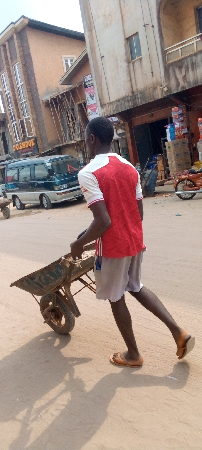 A dark-skinned male individual, viewed from behind, pushes a single-wheeled metal wheelbarrow across a dusty, light-brown road in Nnewi, Nigeria. He wears a red and white patterned short-sleeved jersey, light-colored shorts, and orange flip-flops. His body is angled forward, indicating active movement. Long shadows of the man and wheelbarrow are cast on the road.
In the background, a dark green over silver Mitsubishi van, bearing a partial Nigerian license plate "EML-796 NDA" and a red triangular warning sign on its roof, is parked on the left side of the road. Further right, a reddish-orange motorcycle is visible near a store entrance where various boxed goods are stacked. An empty dark blue wheelbarrow is also present.
Two multi-story buildings line the street. The building on the left features a tan facade with brick-like detailing and multiple white-framed windows. A sign for "D.O. EMDOK INDUSTRIES LTD." is affixed to its lower level. Adjacent to this, on the right, is an older, weathered multi-story building with a lighter-colored concrete facade. Wooden scaffolding is visible in front of or adjacent to this structure. A sign for "T.BAO. RAZORLINE IMPORTER & GENERAL MERCHANT" is also visible on this building. The scene depicts a commercial or mixed-use area.
