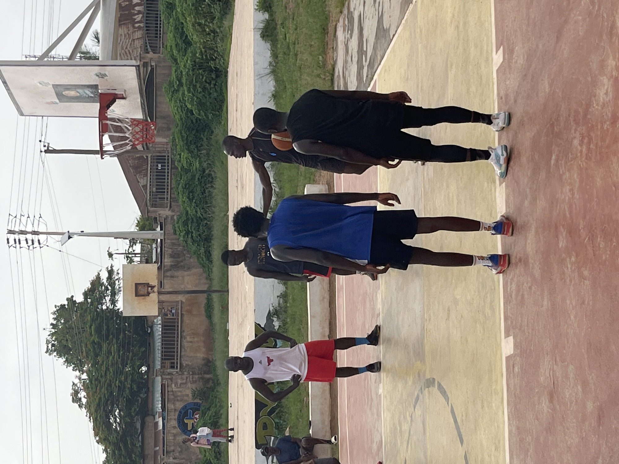 The image shows an outdoor basketball court. Several people are present on the court, some playing and others observing. In the foreground, two individuals are facing away from the camera, wearing basketball attire. One is in a blue jersey and black shorts, and the other is in a black sleeveless shirt and black leggings. Further back, a man in a white tank top and red shorts stands with his hands on his hips, looking towards the center of the court. Another man in a dark grey tank top is holding a basketball and gesturing with his left arm. A man in a dark blue t-shirt and dark shorts sits on a low wall to the left. In the background, there are buildings, trees, and utility poles, suggesting an urban or semi-urban environment. The basketball court itself has a reddish-brown and yellow surface with painted markings. The sky is overcast. Based on the provided information, this scene is located in the city of Gura Top, Nigeria.