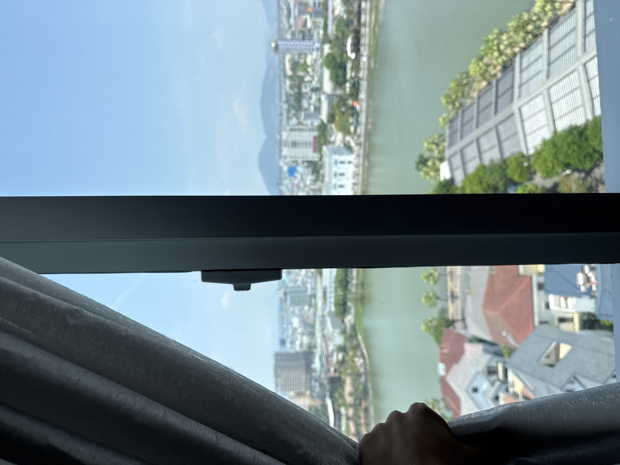 The image presents a view from inside a building, looking out over a city landscape bisected by a river. The focus is on Đà Nẵng, Vietnam. The foreground includes a partially drawn curtain and a hand pulling it back, revealing the exterior view. A window frame is visible, separating the indoor and outdoor spaces.
The river runs through the city, with buildings and vegetation lining its banks. Skyscrapers and residential structures are visible, indicating a densely populated urban area. In the distance, mountains are visible against a clear, blue sky. The river reflects light, suggesting a sunny day. The buildings nearest to the vantage point appear to be residential, with visible roof structures. The overall scene suggests a mix of urban development and natural landscape in Đà Nẵng.