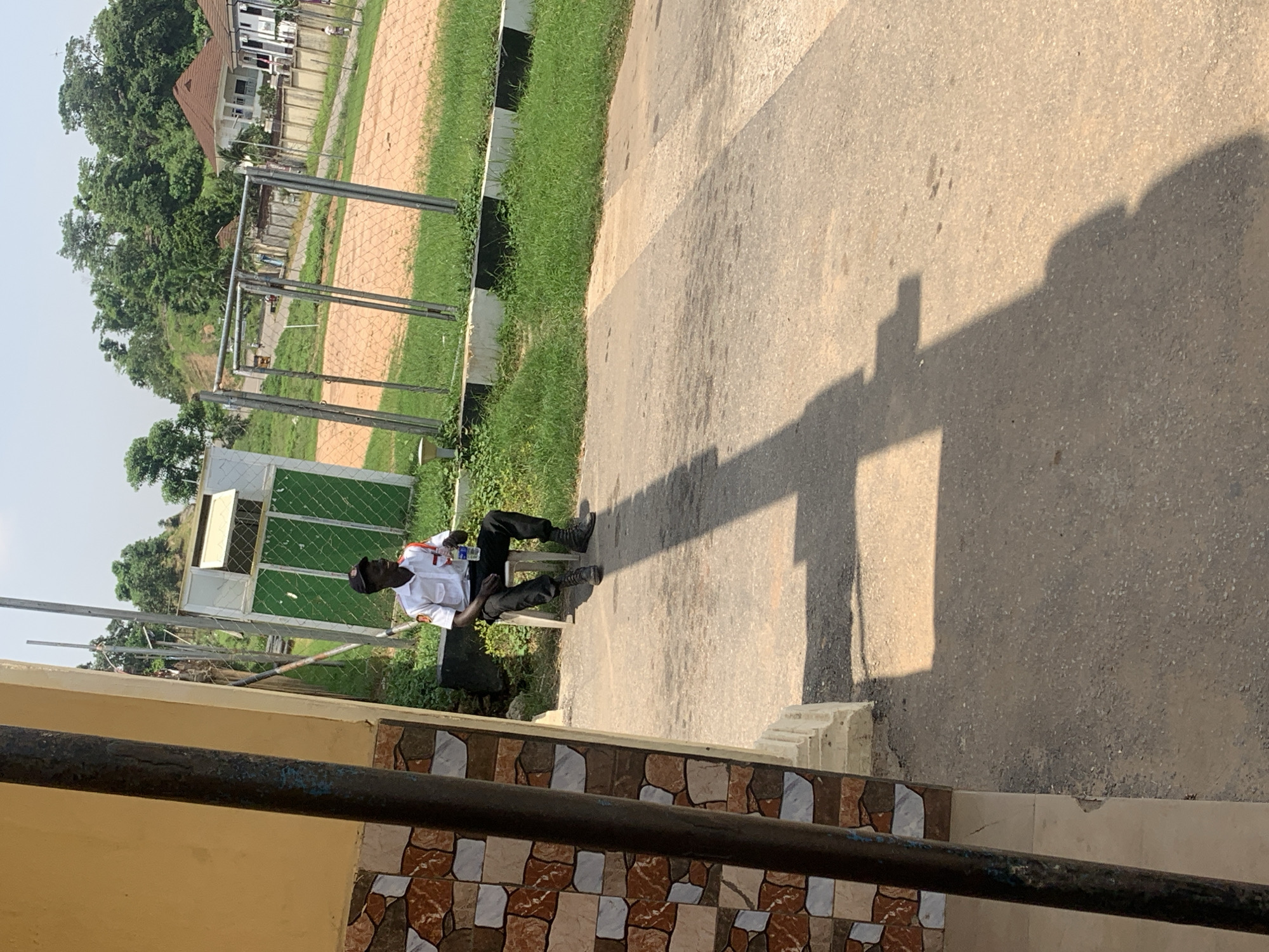 A person is sitting on a white chair in front of a building. The person is wearing a white shirt with red trim, black pants, and black boots. The person is holding a bottle of water. The building has a yellow facade and a tiled wall. In the background, there is a green fence, a sandy field, and some houses on a hill. The image appears to be taken in Abuja, Nigeria, given the context of the location. The lighting suggests it is daytime.