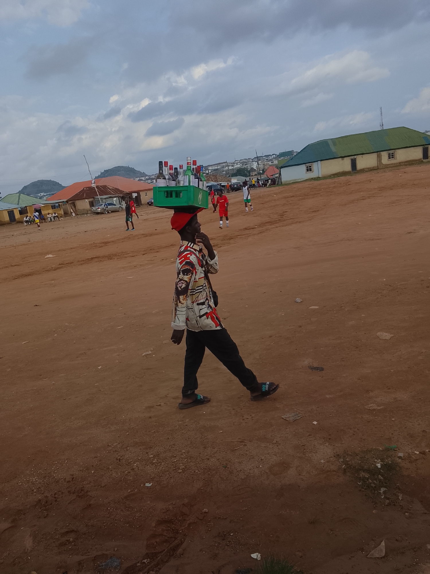 A young man walks across a dusty, undeveloped area in Abuja, Nigeria, carrying a green crate filled with bottles on his head. He is wearing a patterned long-sleeved shirt and black pants. In the background, there are several low-rise buildings with green roofs and some scattered people, some of whom appear to be playing a sport. The sky is cloudy and overcast. The scene suggests a casual outdoor gathering or market in a less developed part of the city.