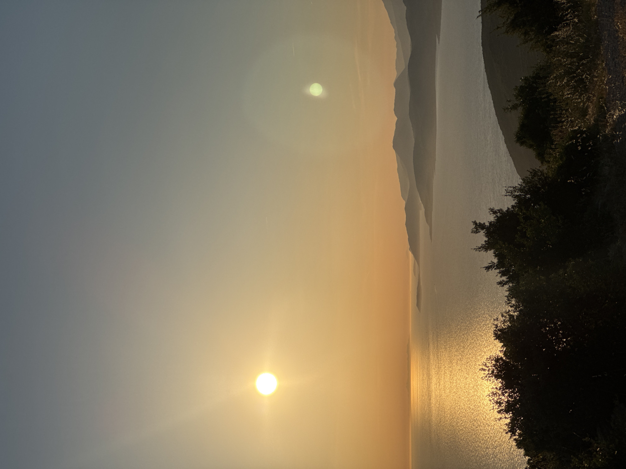 The image depicts a sunset over the sea, as viewed from a hilltop overlooking Kovačko Polje, Montenegro. The sun, a bright, glowing orb, is positioned in the upper left quadrant of the frame, casting a golden sheen across the water. The sea itself is calm, reflecting the warm hues of the sunset, with distant, silhouetted mountains forming a layered backdrop along the coastline. In the foreground, the dark silhouettes of trees and vegetation create a stark contrast against the luminous sky and sea, suggesting a viewpoint from a densely vegetated area. The sky transitions from a pale yellow near the horizon to a soft blue at the top of the frame. A second, fainter circular lens flare is visible in the upper right portion of the sky. There are no people or animals visible in the image. The overall scene is serene and natural, dominated by the elements of sky, sea, and land at dusk.