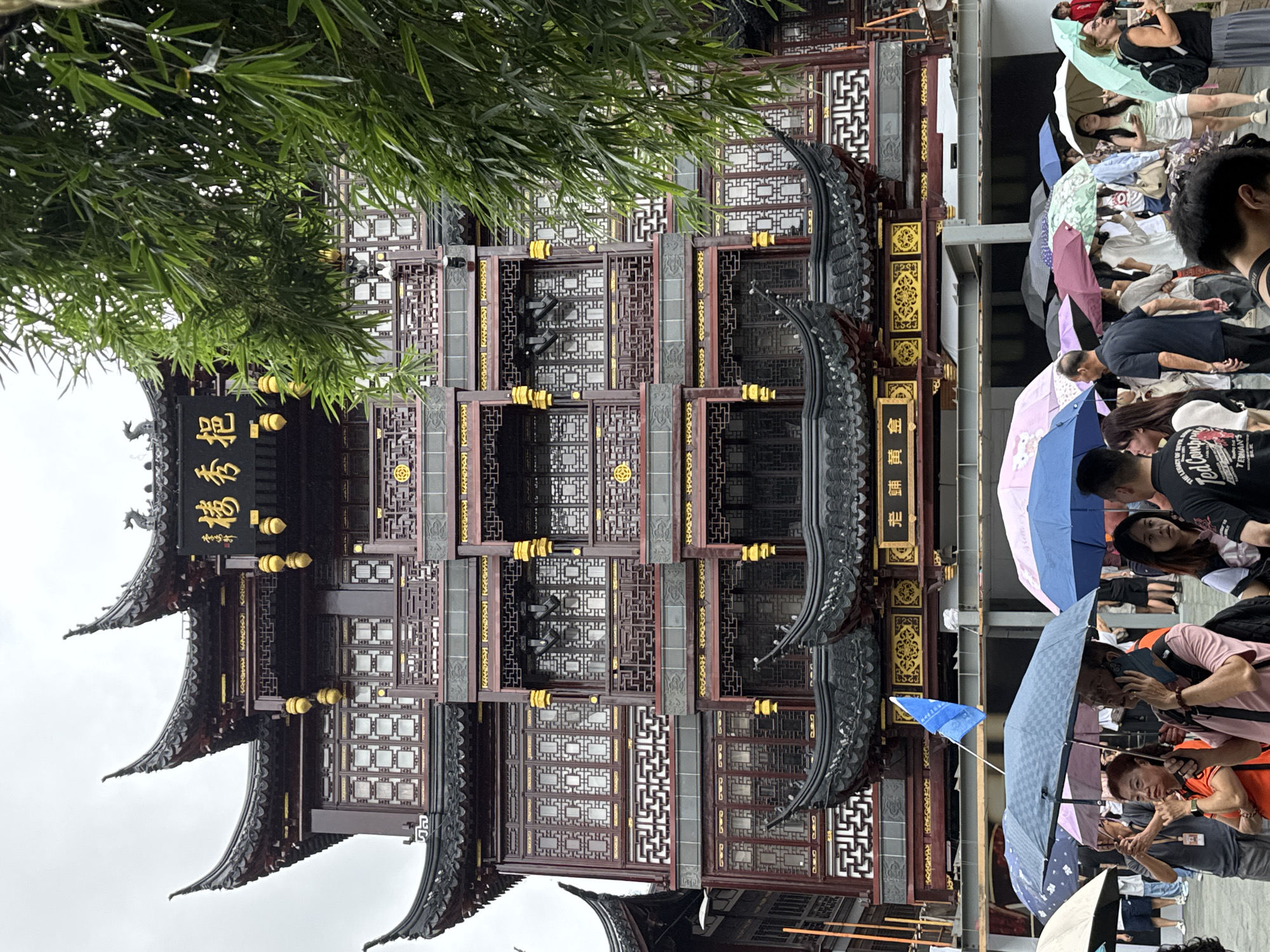 The image shows a view of the Yu Garden Bazaar in Shanghai, China, on a cloudy day. In the foreground, a crowd of people are holding umbrellas, indicating rain or strong sun. The people are diverse in age and gender, and are dressed casually. Some are walking, others are talking on their phones or looking at something. Behind the crowd, a multi-story, traditional Chinese building with ornate architecture, including curved roofs and intricate wooden carvings, dominates the scene. The building is decorated with gold accents and Chinese characters. A few trees with green leaves are visible on the right side of the image, partially obscuring the building. A blue flag is also visible, attached to a pole in front of the building. The overall atmosphere suggests a tourist area.